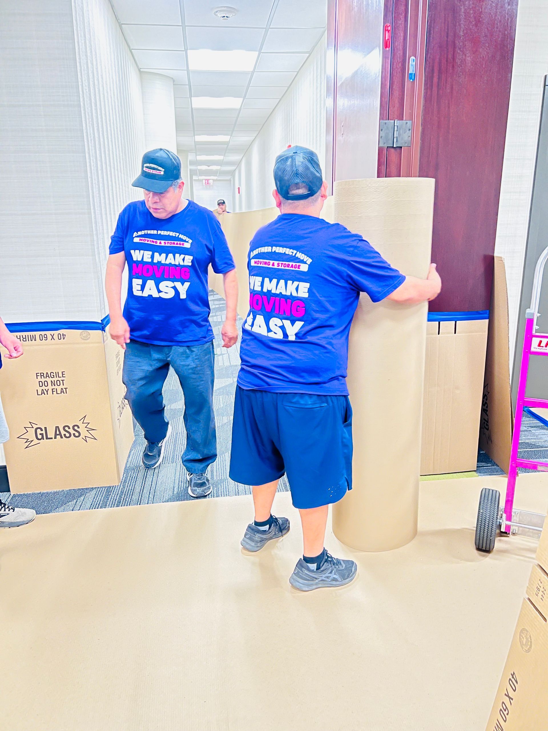 Two men are carrying large rolls of cardboard in a hallway.