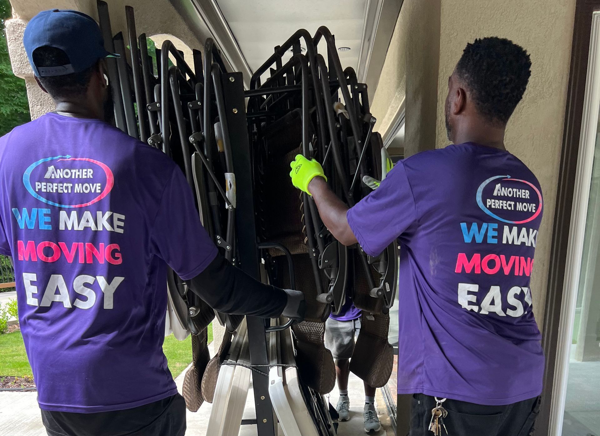 Two men wearing purple shirts that say `` we make moving easy '' are carrying a stack of chairs.