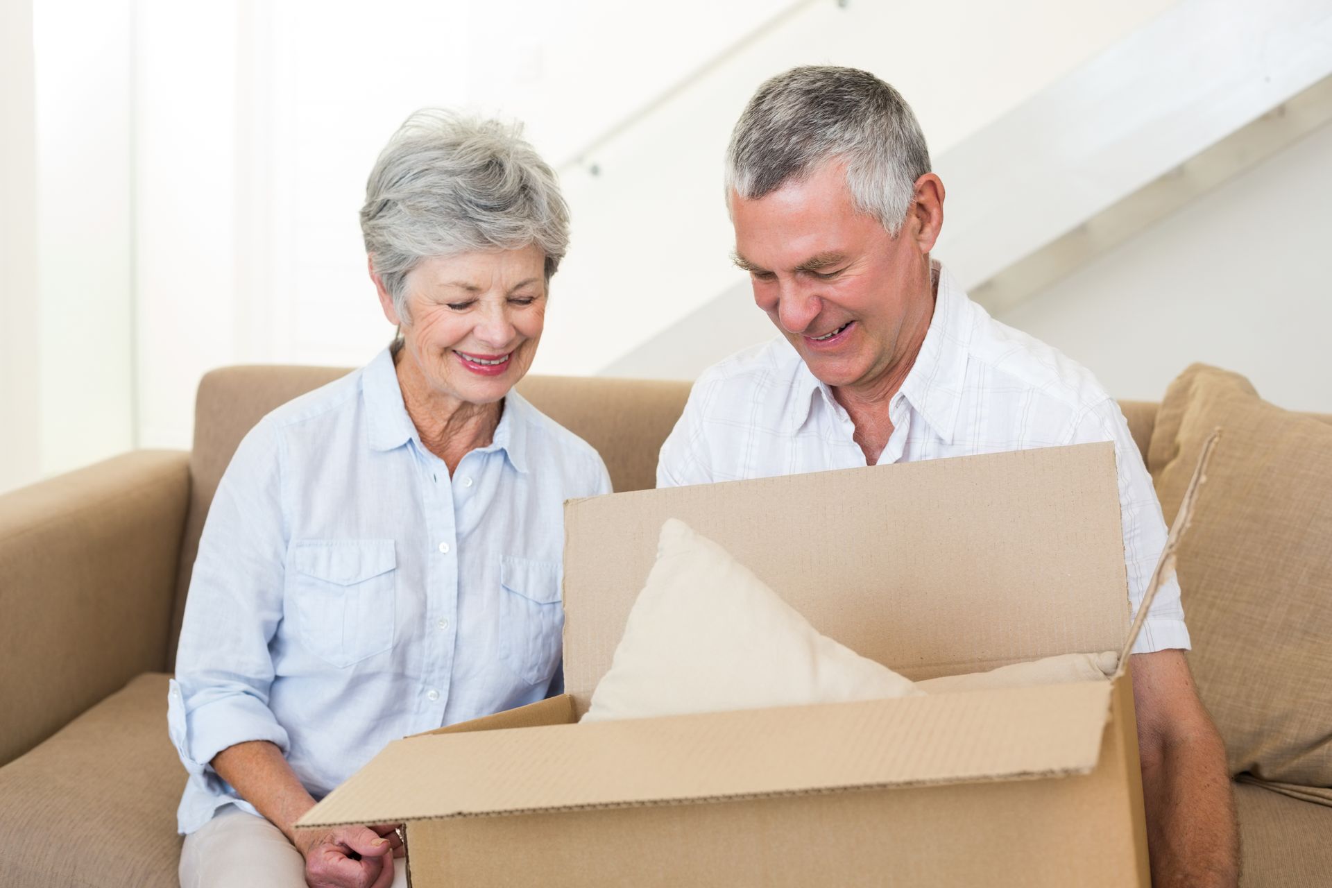 An elderly couple is sitting on a couch opening a cardboard box.