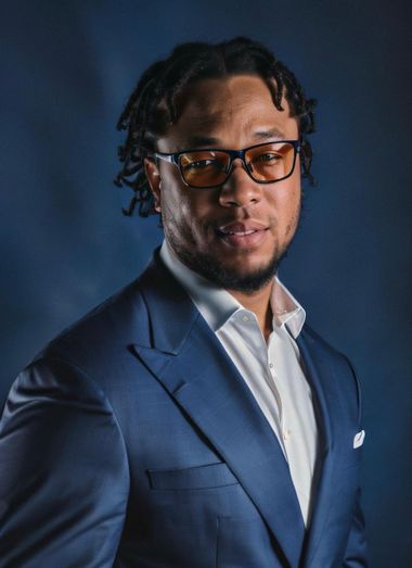 A man in a suit and glasses is standing in front of a blue background.