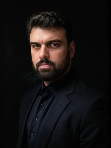 A man with a beard is wearing a black suit and a black shirt.