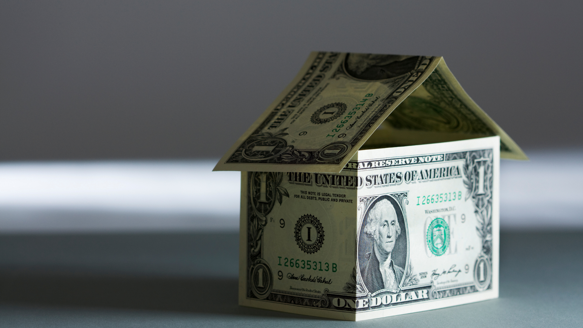 A small house made of US dollar bills, casting a shadow on a light gray surface.