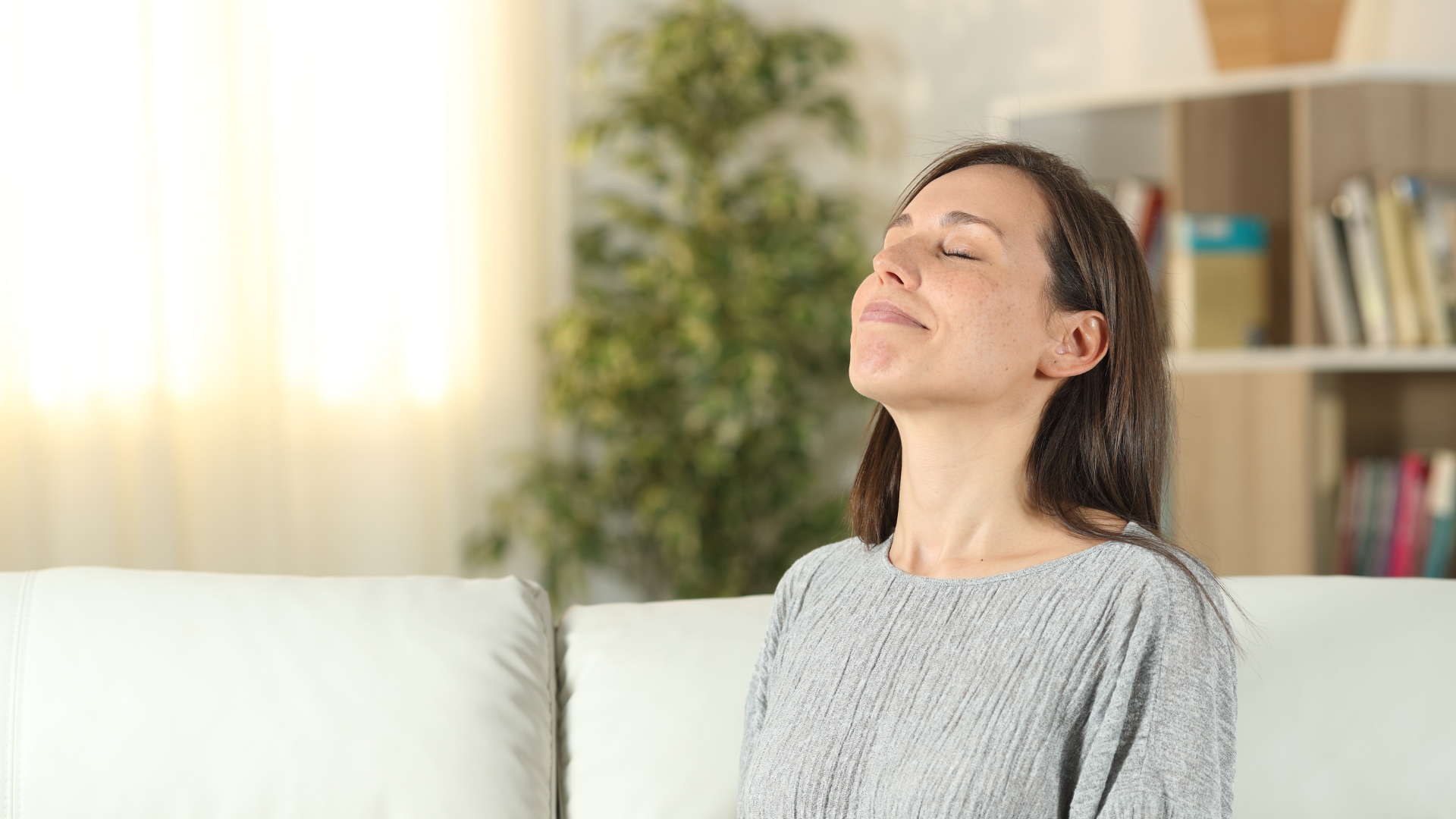 Woman with eyes closed, taking a deep breath on a white couch, with a plant and bookshelf in the background.