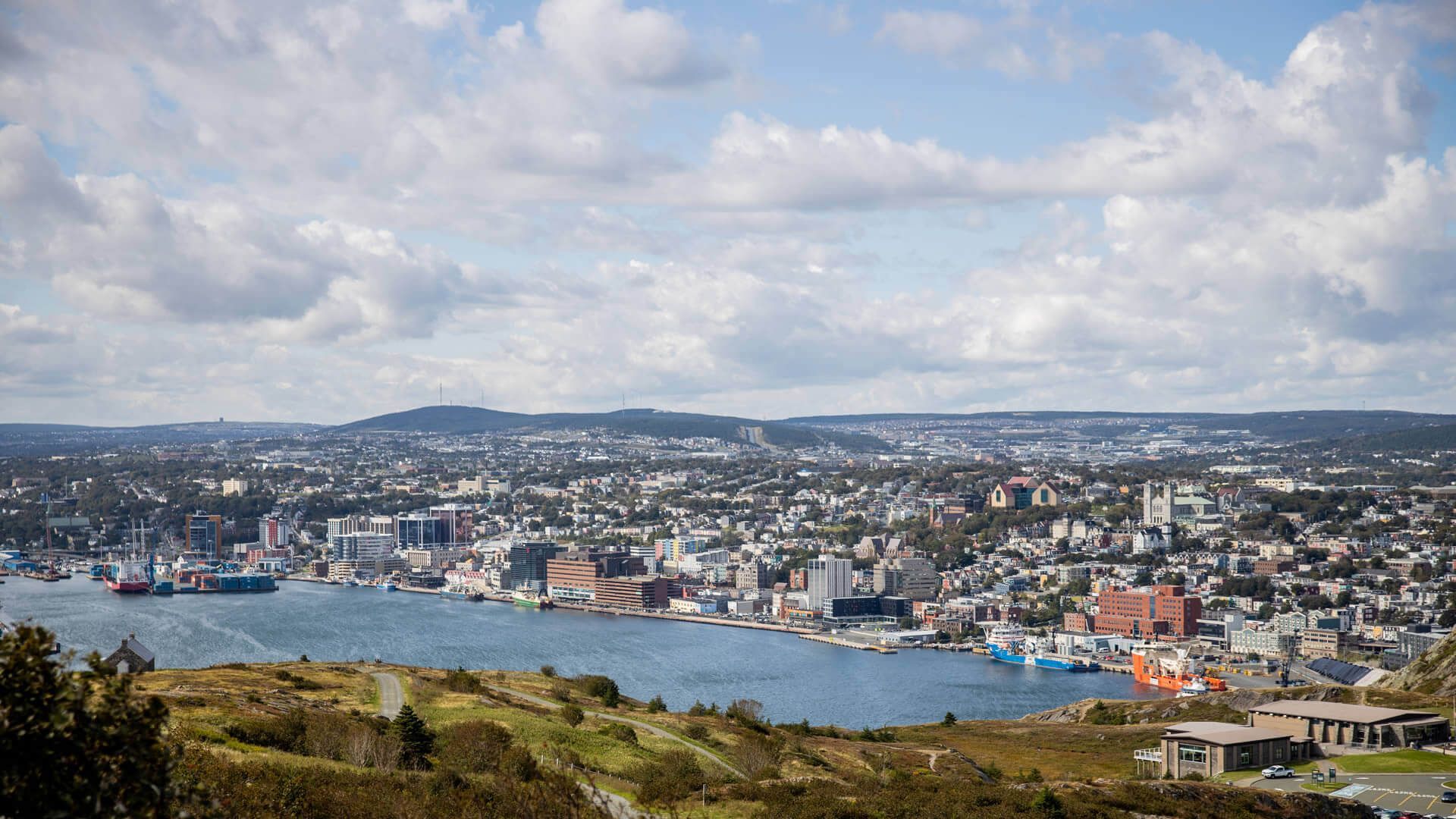 View of a coastal city with harbor, buildings, and hills under a partly cloudy sky.