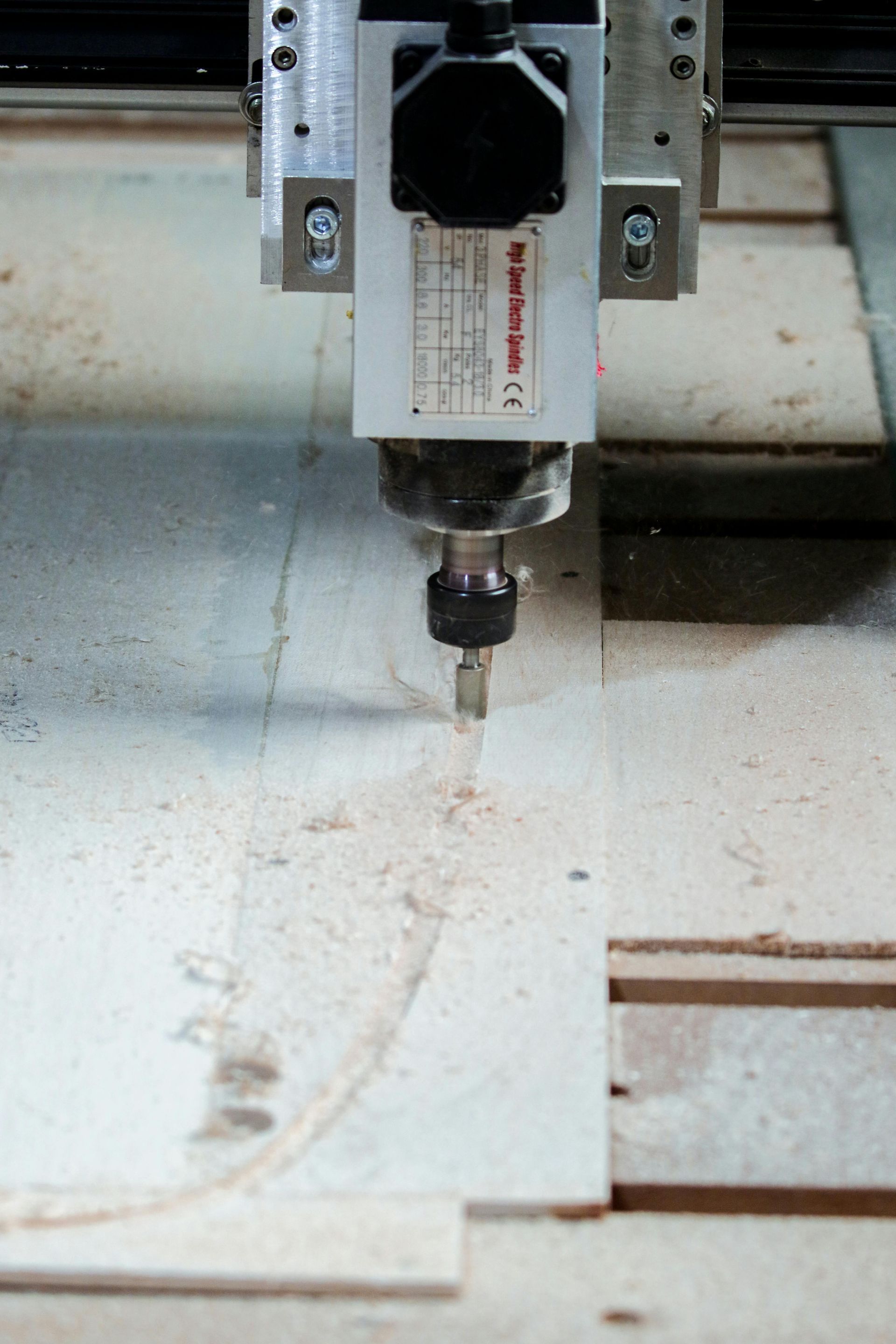 CNC router cutting wood, sawdust visible. Close-up of cutting bit and machine detail.