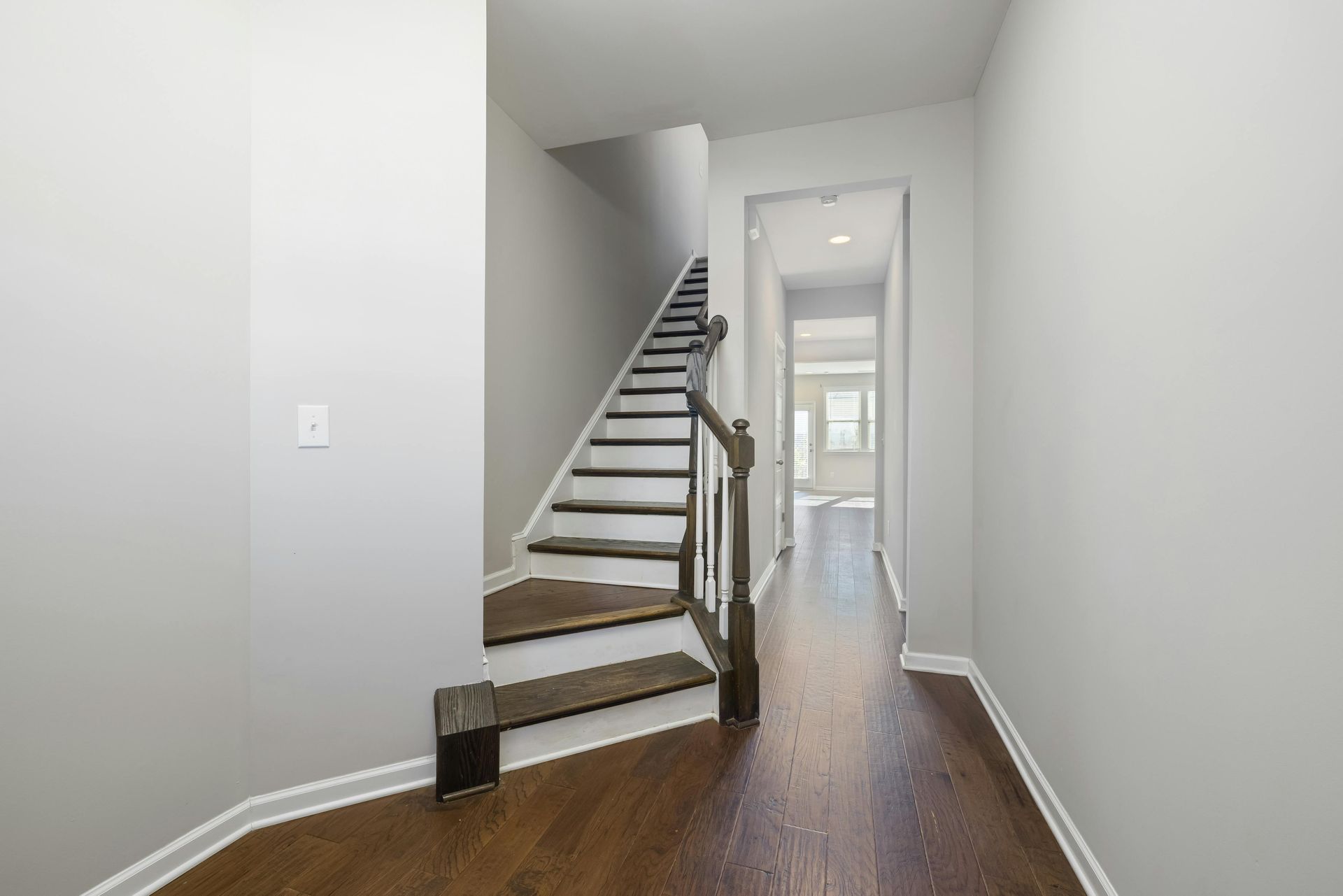 Hallway with staircase and hardwood floors, leading to a doorway. Walls are gray.