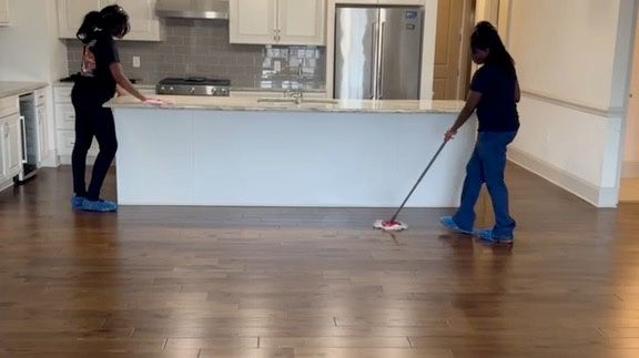 Two people cleaning a hardwood floor in a kitchen: one leaning on a counter, the other mopping.