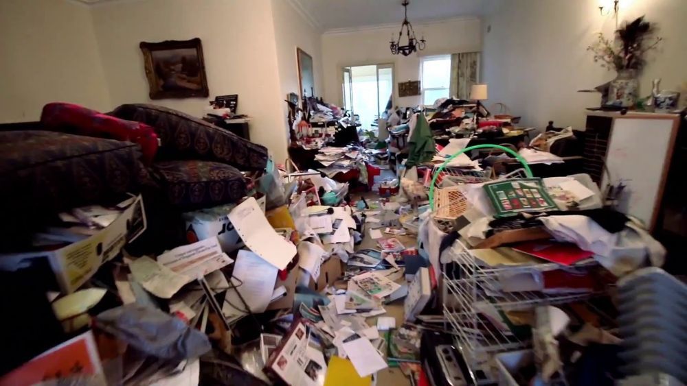 Messy living room filled with piles of papers, books, and clutter across the floor and furniture