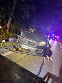 Car accident scene: Gray SUV with front-end damage on a dark, wet road. Police lights flashing in background.