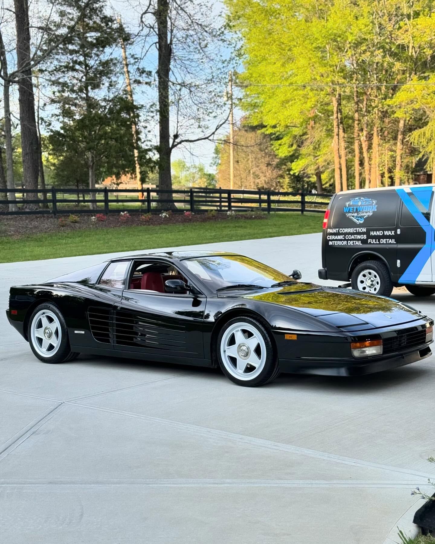 Black Ferrari Testarossa parked on a driveway with white wheels, red interior, and a service van in the background.