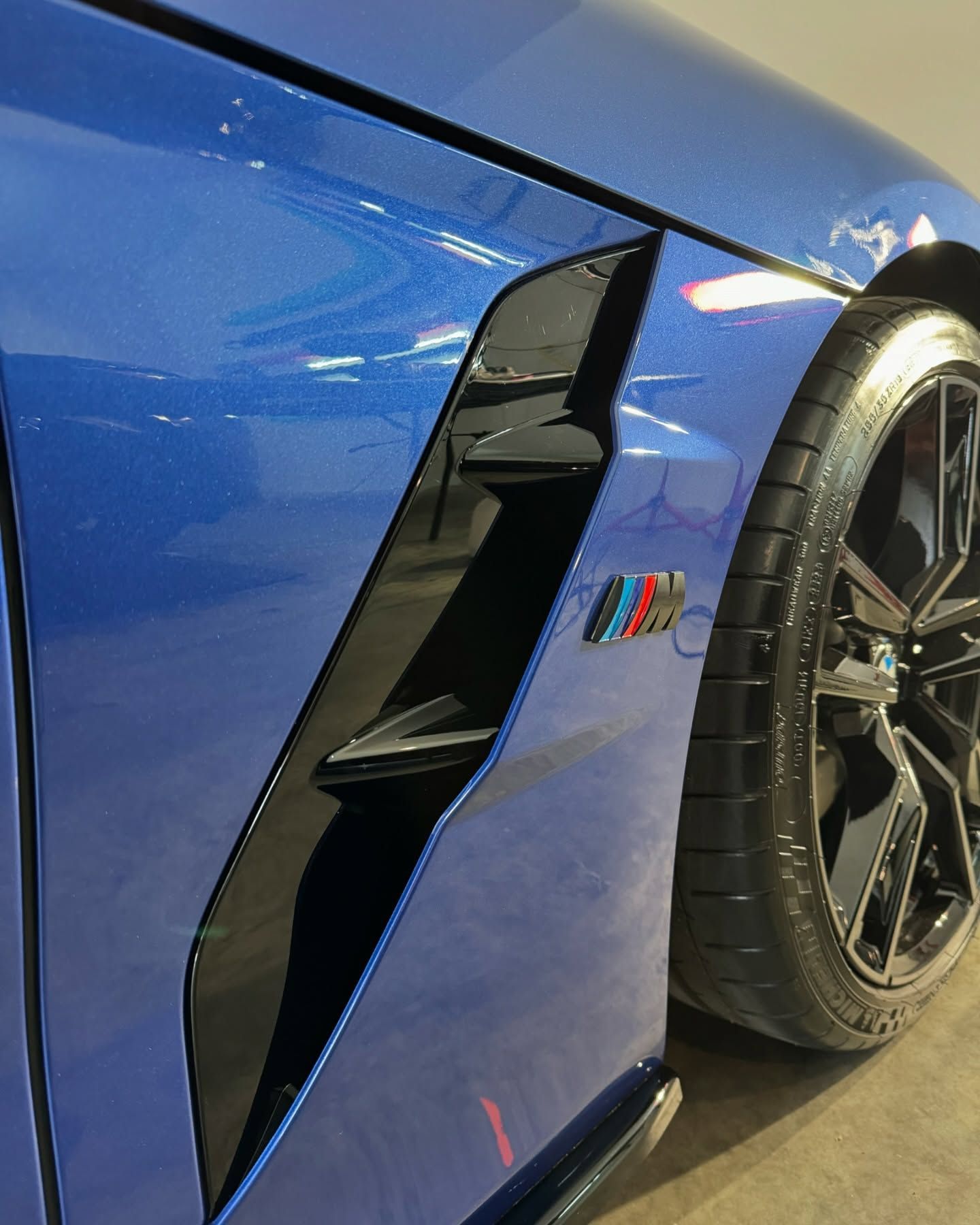 Blue BMW car side detail, air vent, M badge, black wheel and tire.