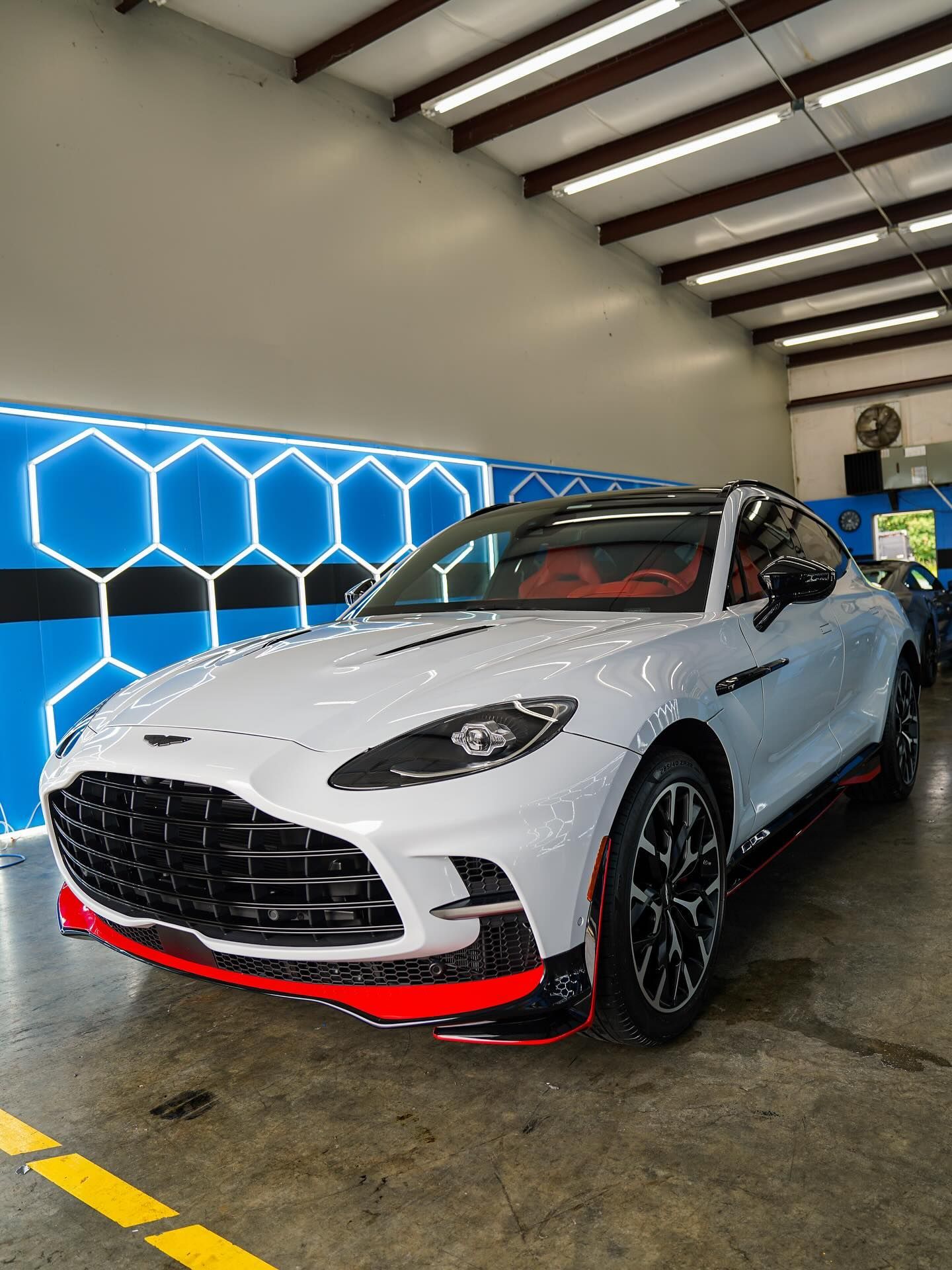 White Aston Martin DBX SUV with red accents parked inside a garage, with blue hexagonal wall decor.