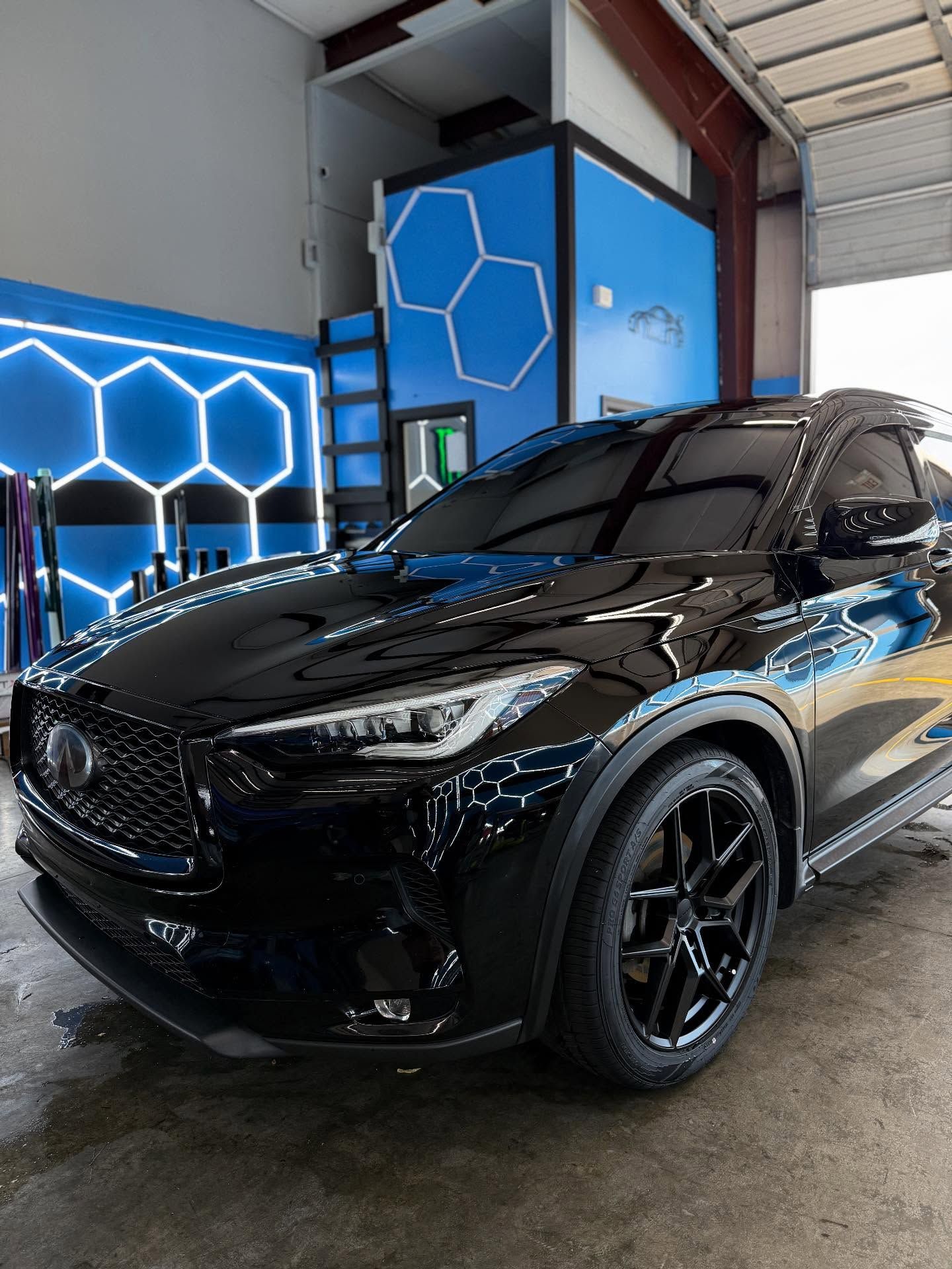 Black Infiniti QX50 SUV with tinted windows, parked in a car detailing bay, with blue hexagonal neon lights.