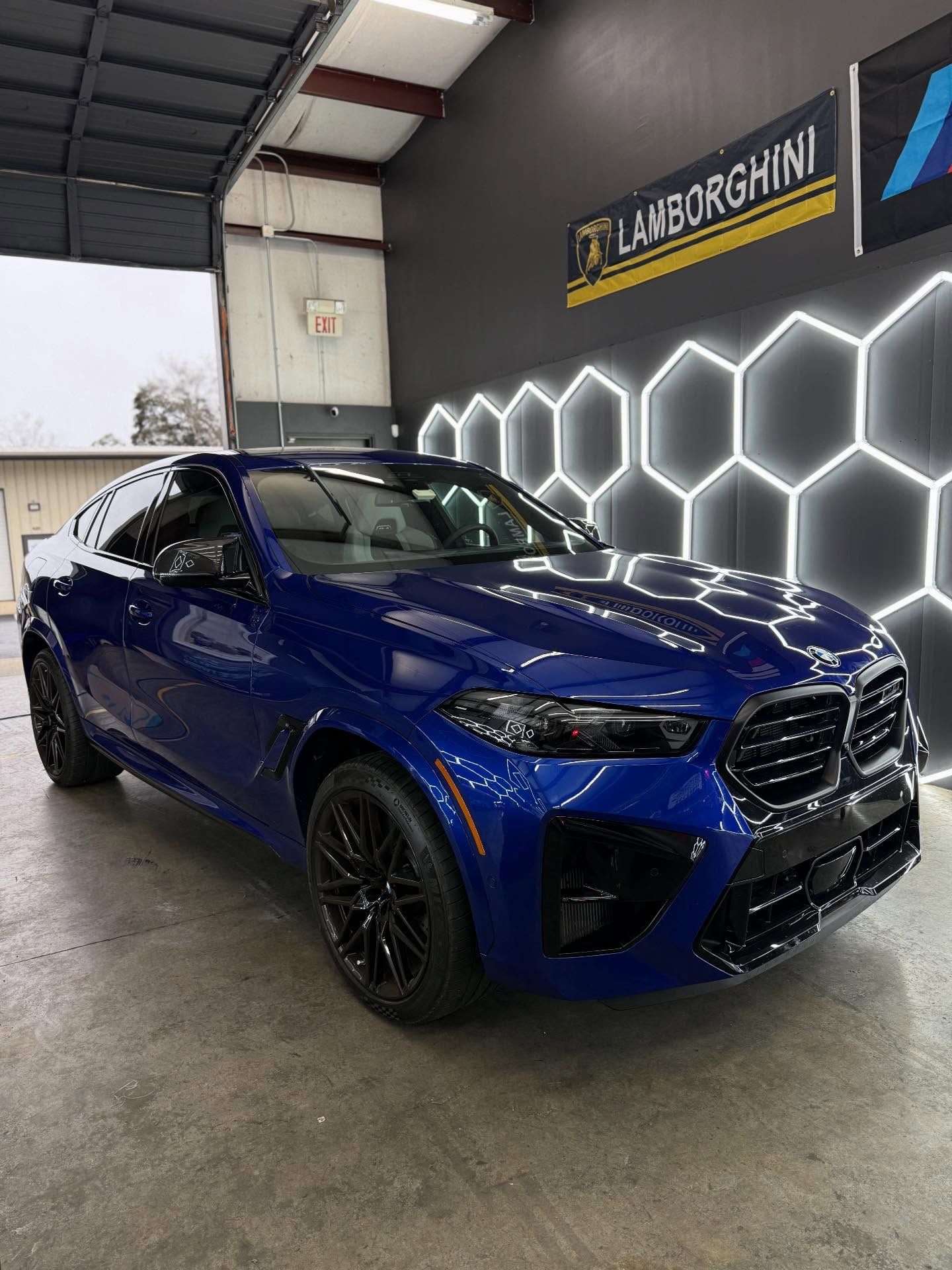 Blue BMW X6 M SUV in a garage with black wheels, honeycomb lighting, and a Lamborghini sign.