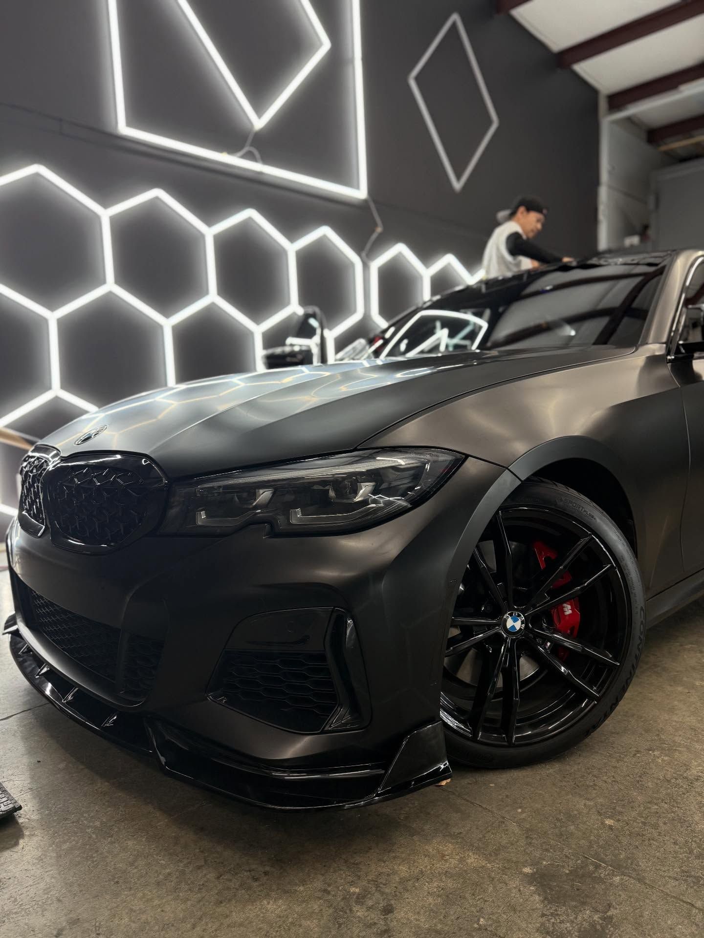 Black car with red brake calipers being worked on in a garage with honeycomb-patterned lights.