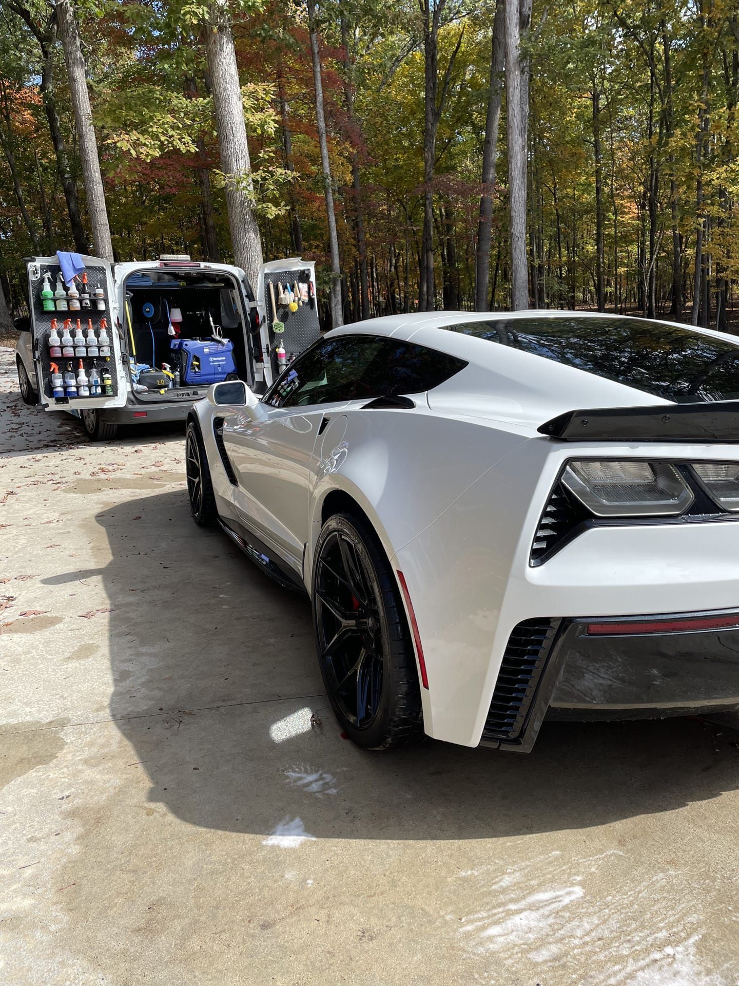 White sports car parked near a van with open doors, possibly detailing service in an outdoor setting with trees.