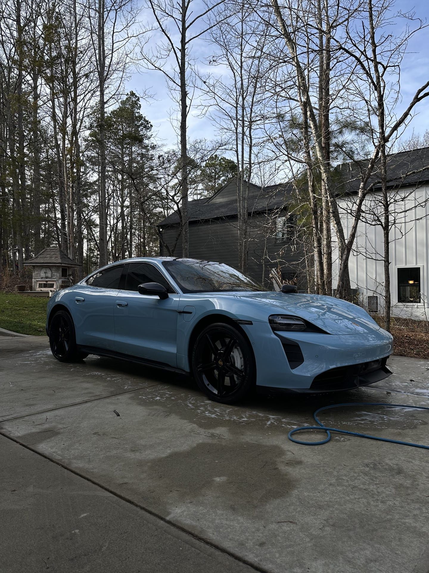 Light blue Porsche Taycan parked on a concrete driveway. Black wheels, trees, and a house in the background.