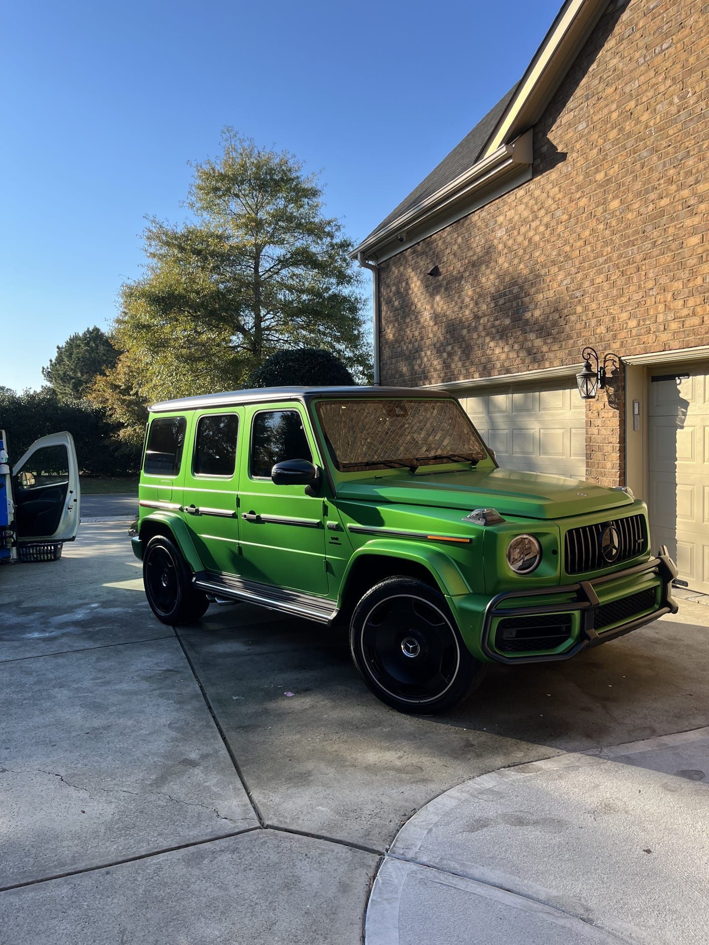 Green Mercedes-Benz G-Wagon parked on a concrete driveway in front of a brick building on a sunny day.