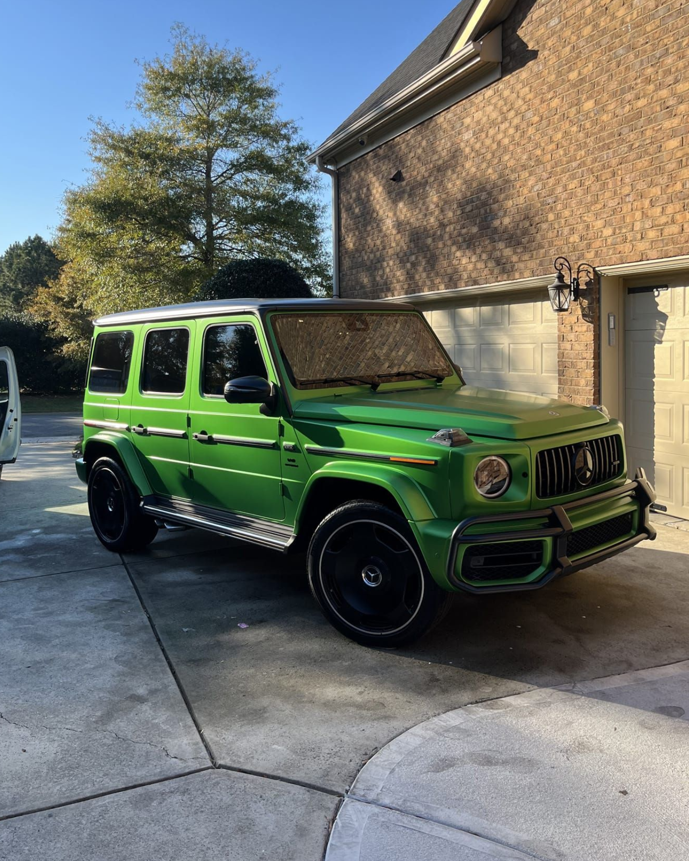 Green Mercedes-Benz G-Wagon parked on a paved driveway in front of a brick building on a sunny day.
