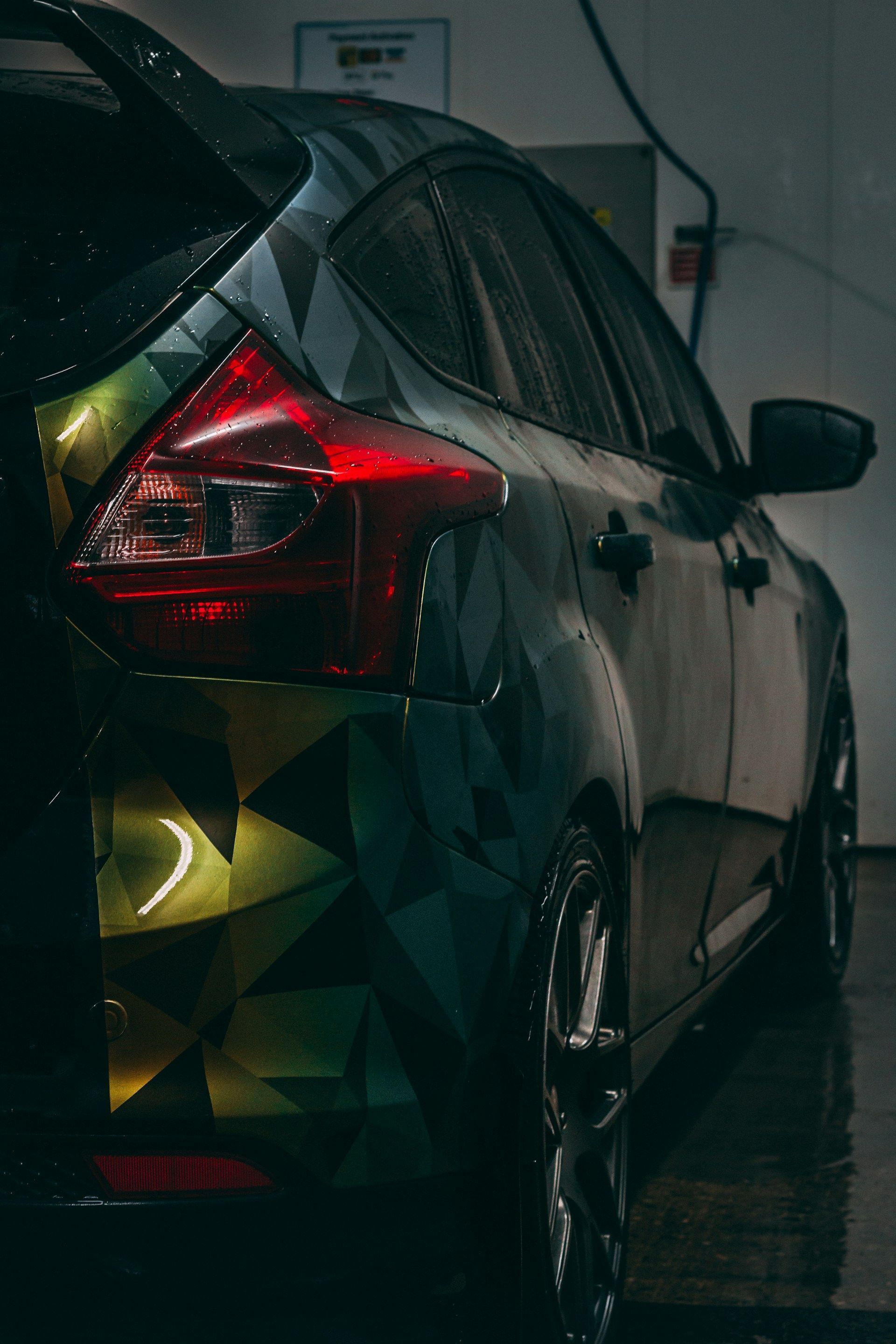 Black car with geometric wrap and illuminated taillight, wet in a wash bay.