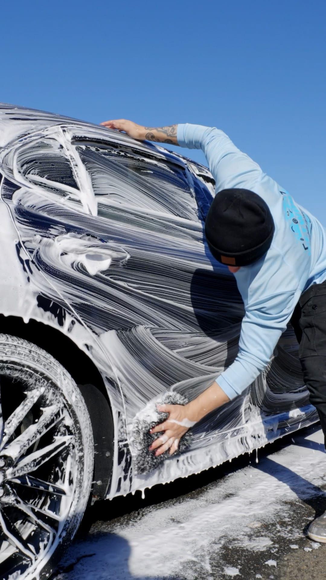 Person washing a black car with soapy water, outdoors on a sunny day.
