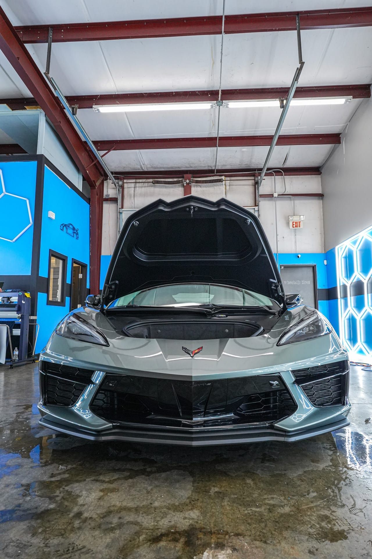 Gray sports car with hood open in a garage, blue walls with honeycomb pattern.