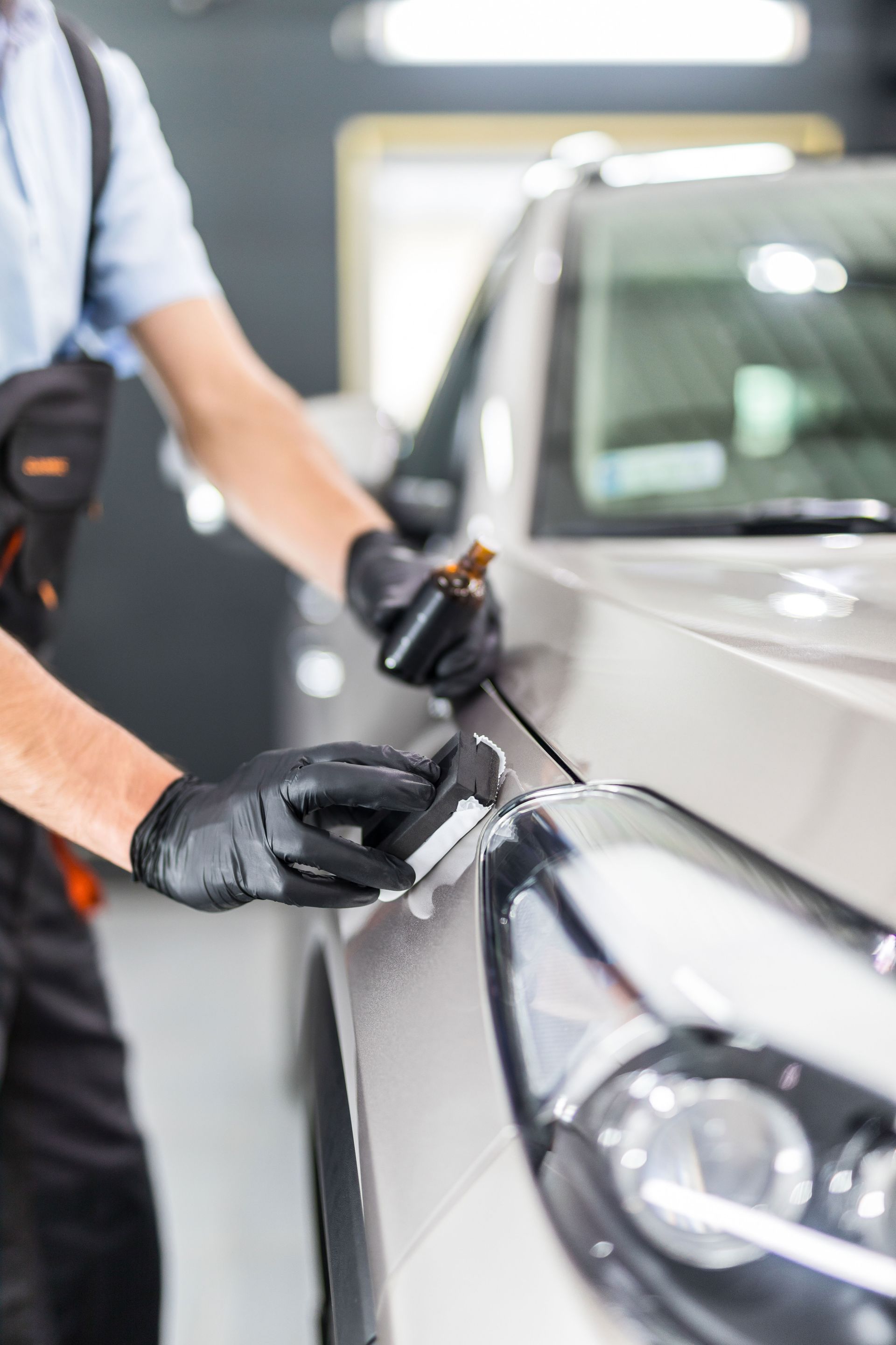 Person applying a coating to a car with black gloves; light car and shop in background.