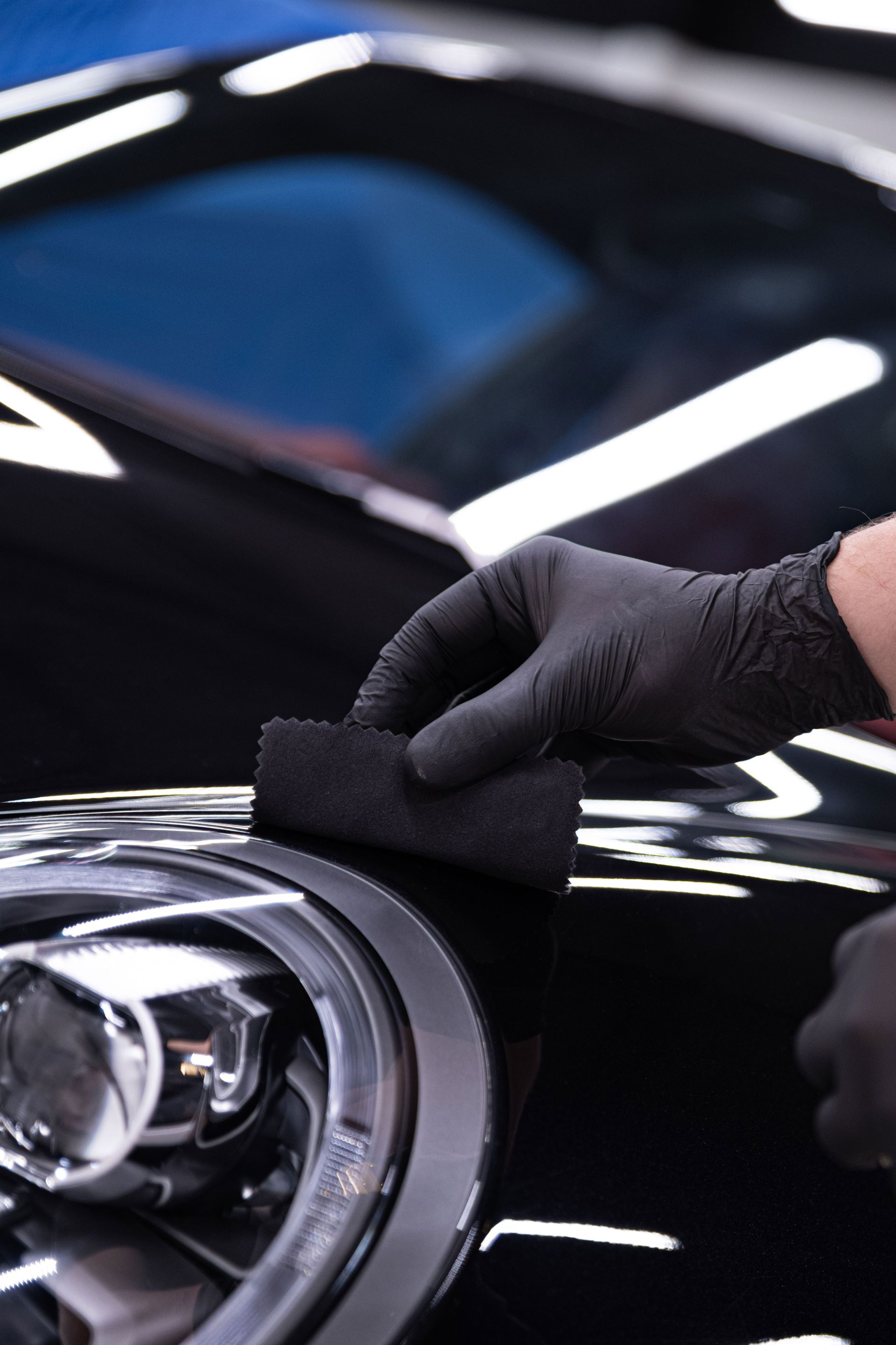 Black-gloved hands applying coating to black car's headlight area with a black applicator in a brightly lit setting.