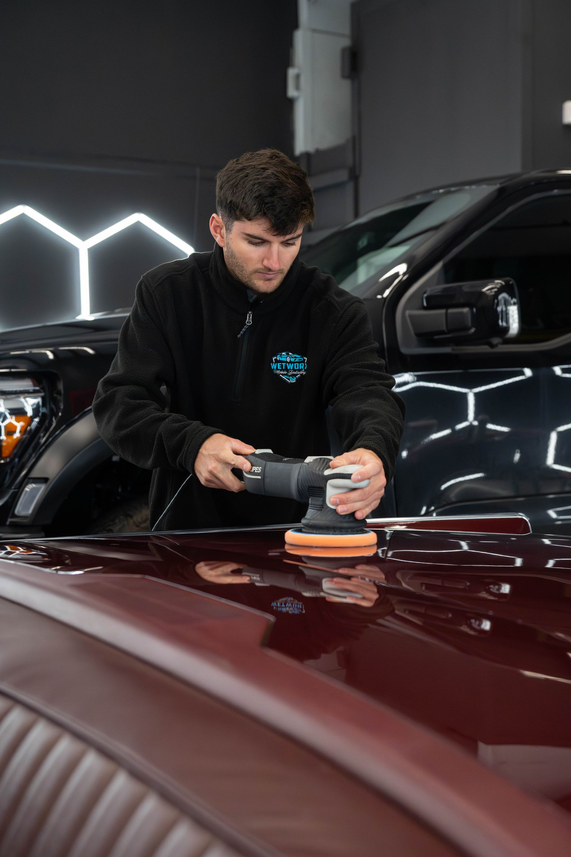 Man polishing a burgundy car hood with a machine in a well-lit shop.