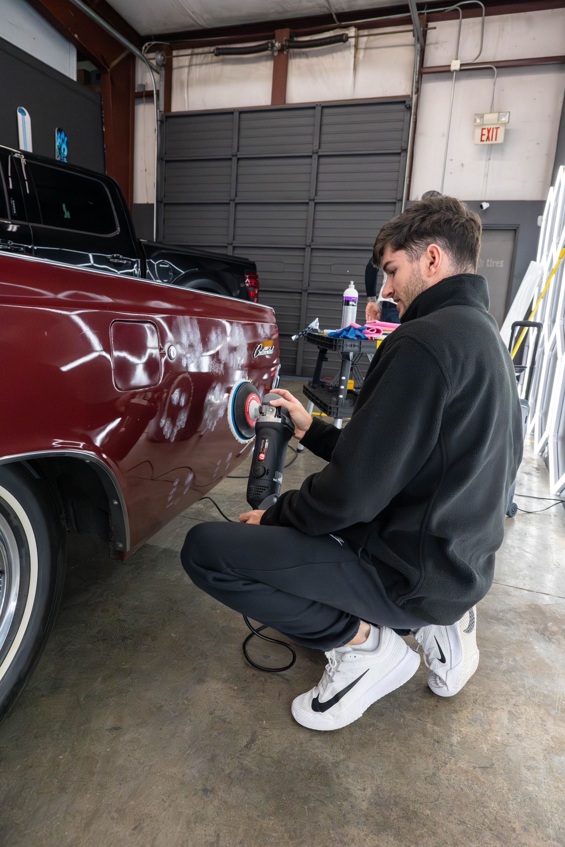 Man polishing a burgundy truck in a garage; he kneels, holding a polisher.