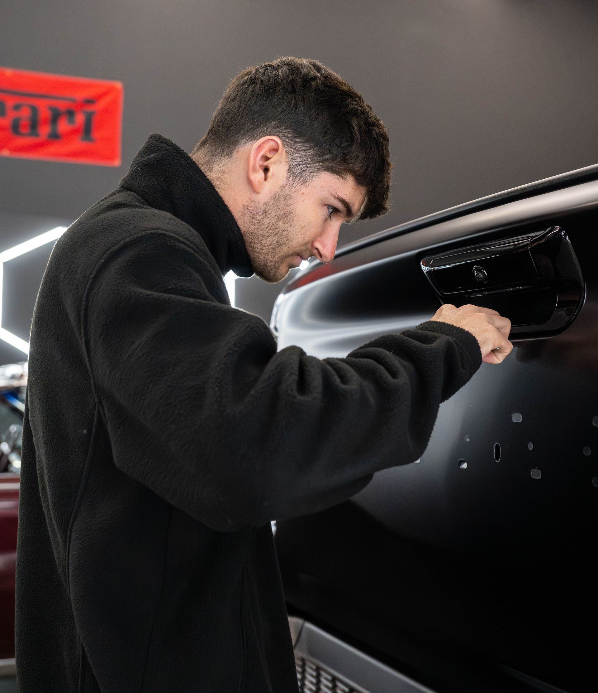 Man in black jacket examining black car door handle in a shop with red Ferrari sign.
