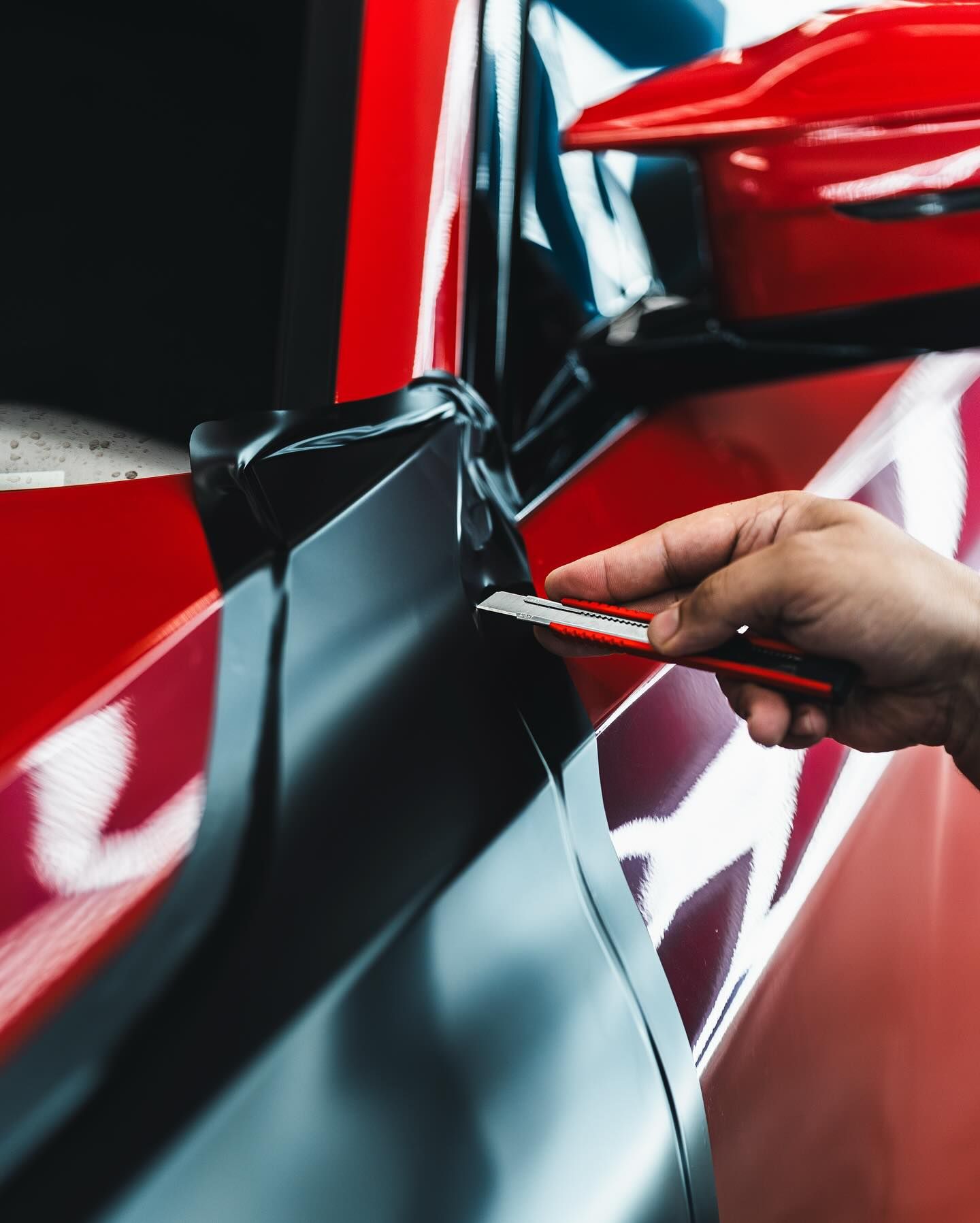 Hand using a blade to trim black vinyl on a red car body panel.