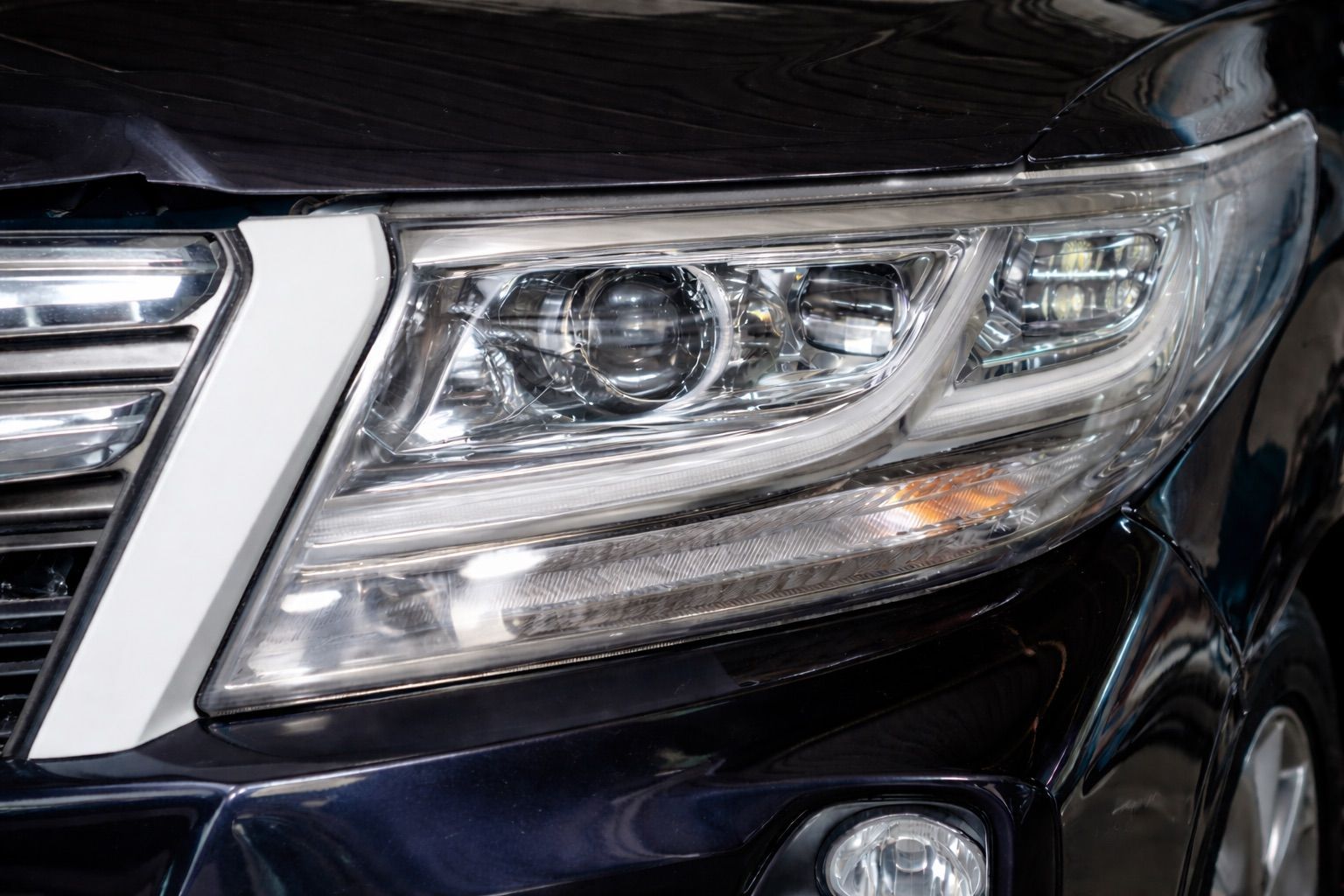 Close-up of a black car's headlight; multiple light sections and chrome trim, against a dark blue car.