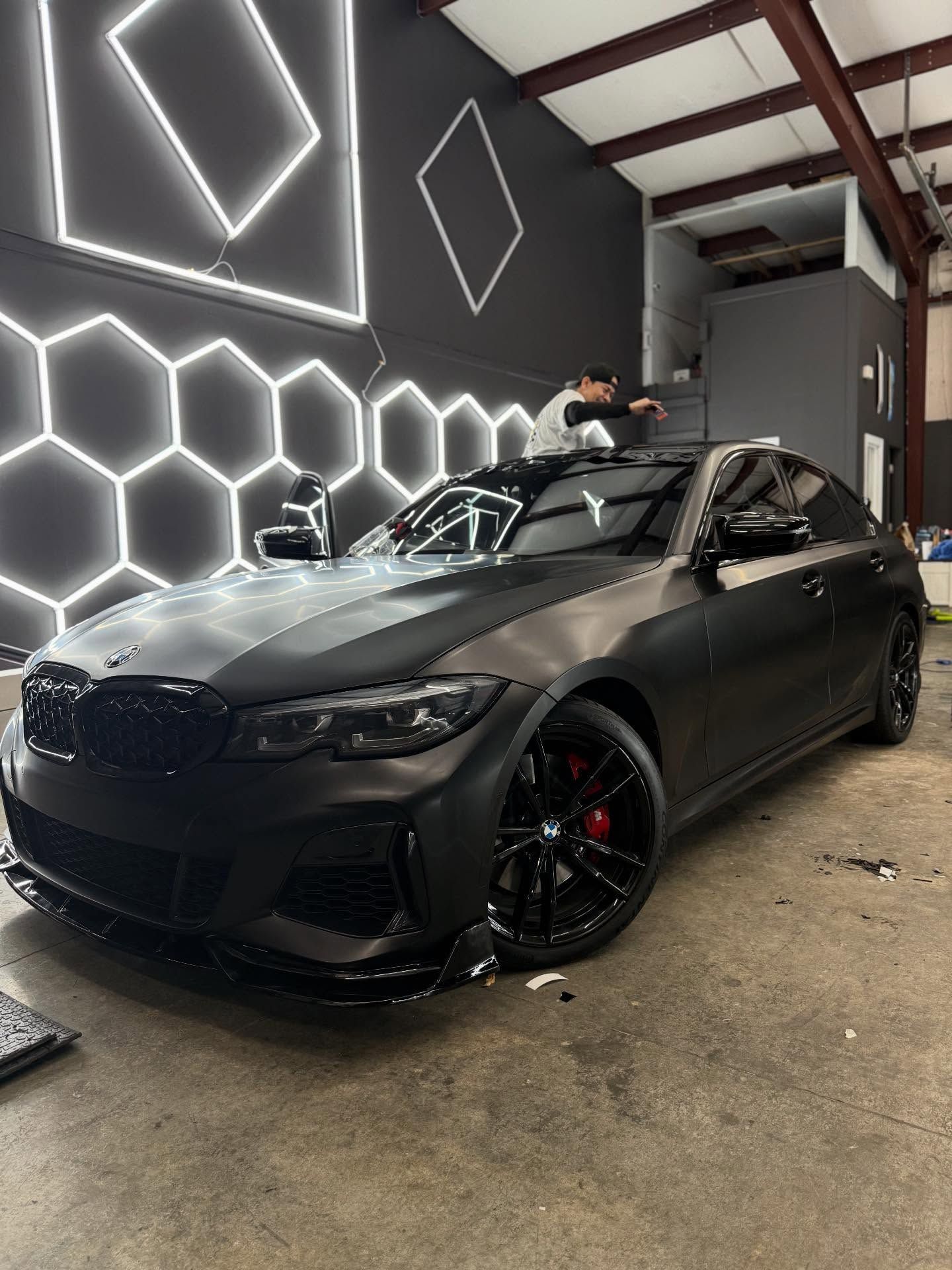 Black BMW sedan in a workshop, being worked on. Red brake calipers, black wheels, and hexagon lights on the wall.