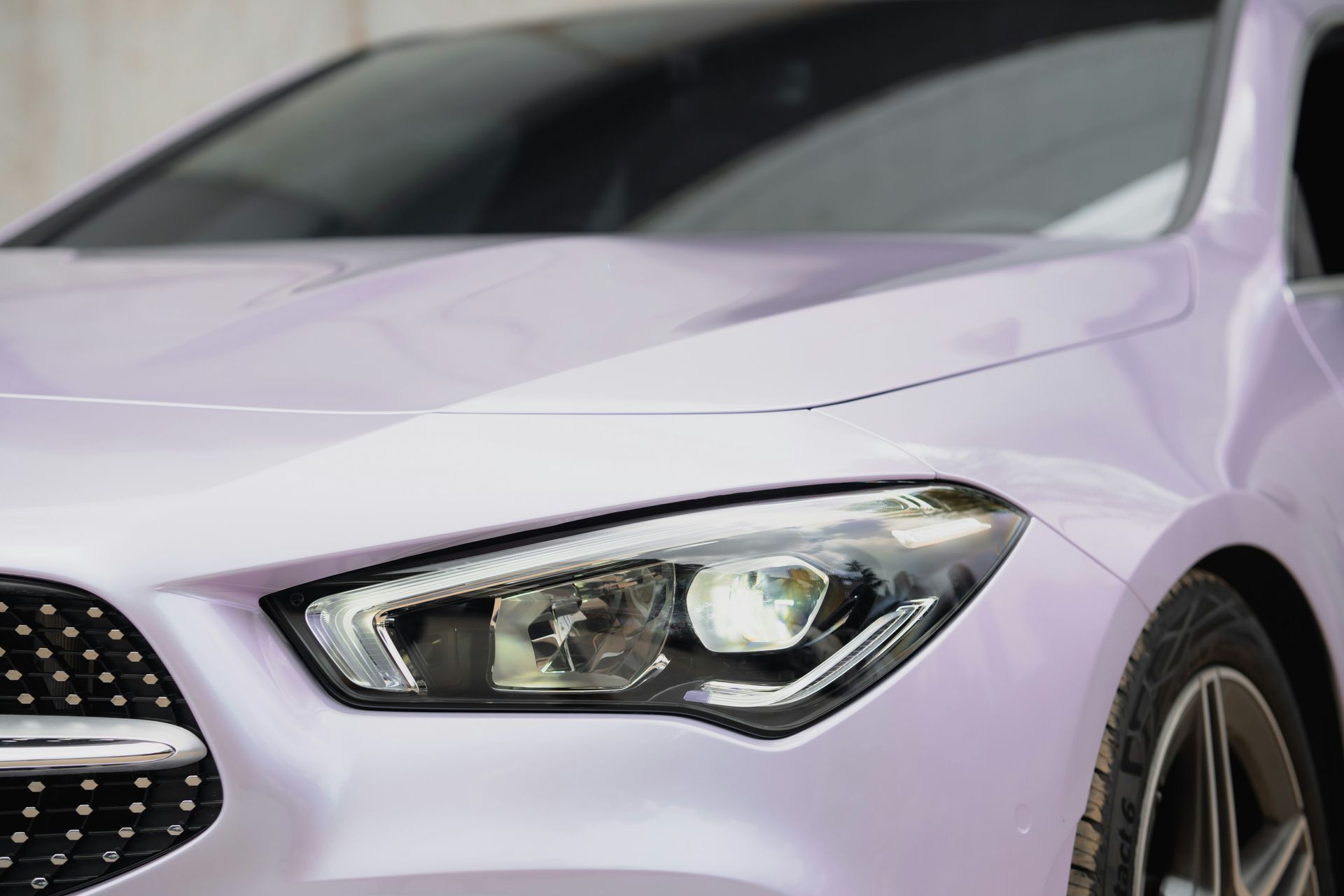 Close-up of a lavender car's front with a lit headlight and black grille.