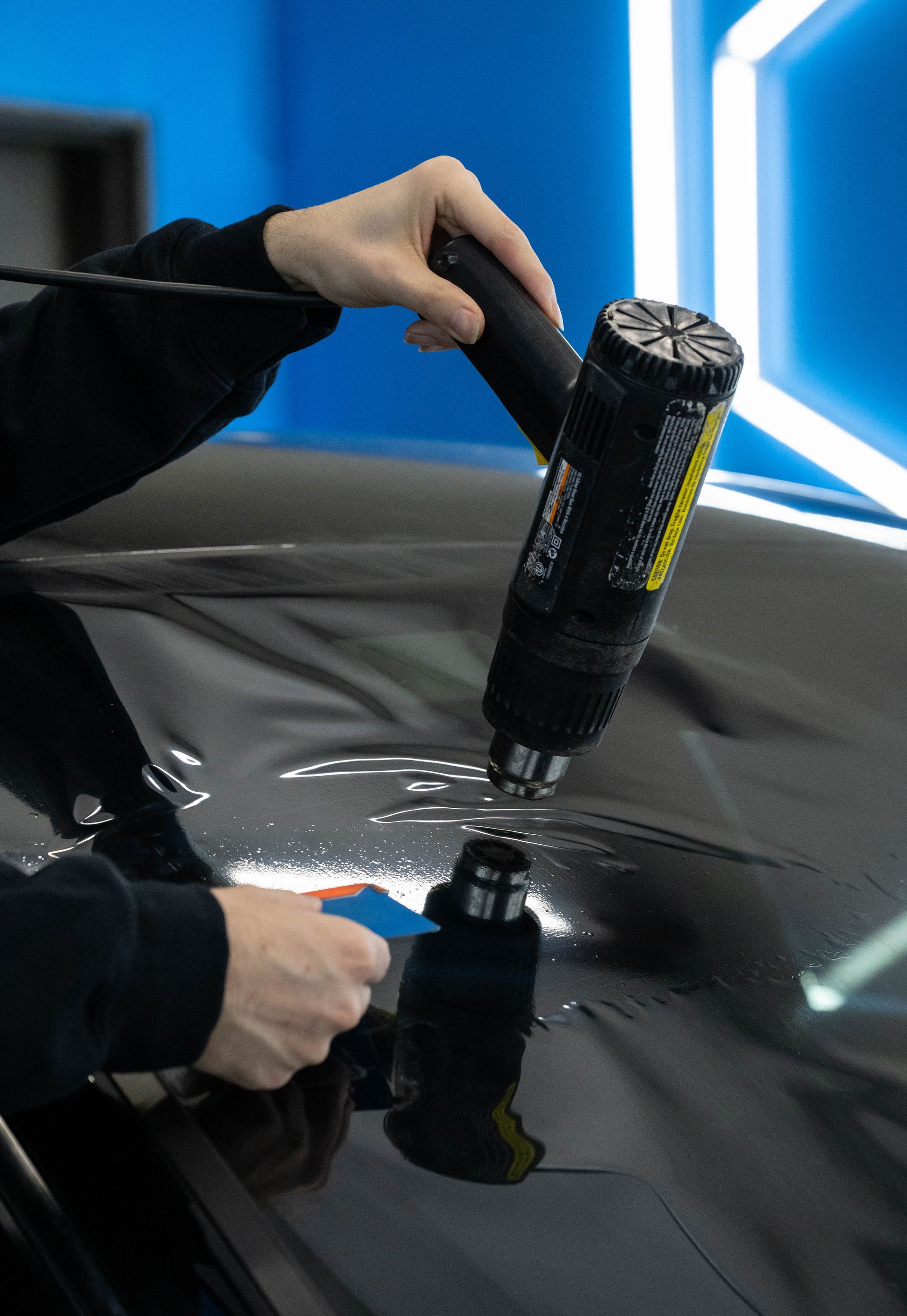 Person using a heat gun to apply film to a vehicle's dark surface in a well-lit workshop.