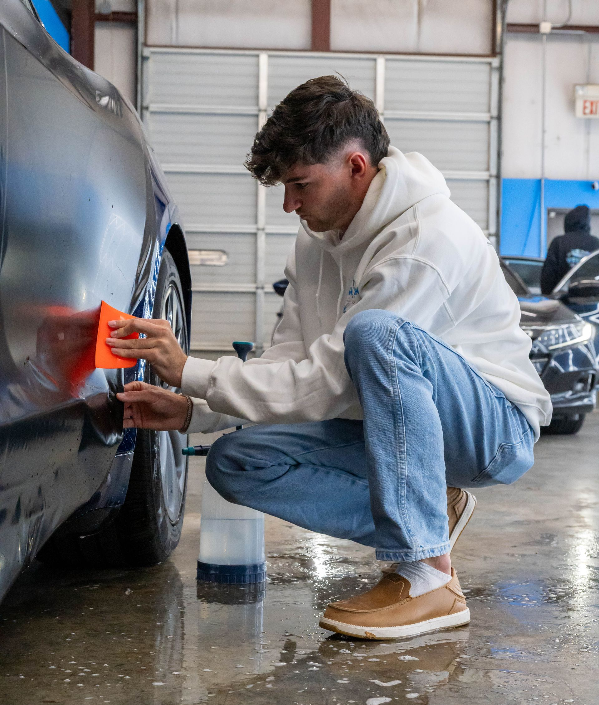 Person washing a car. He is crouching near the wheel with an orange cloth, wearing a white hoodie and jeans.