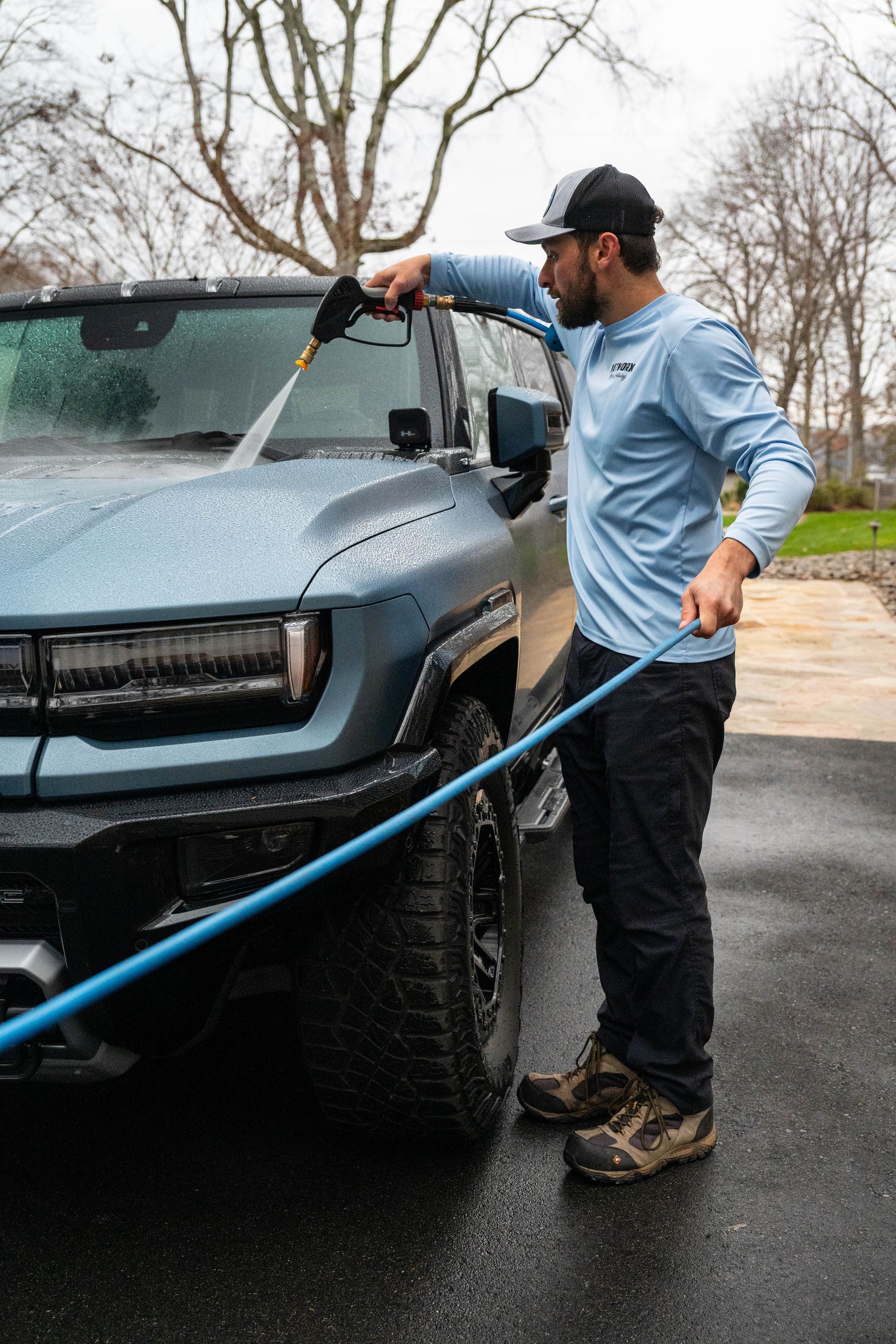 Man washing a blue truck with a pressure washer in a driveway.