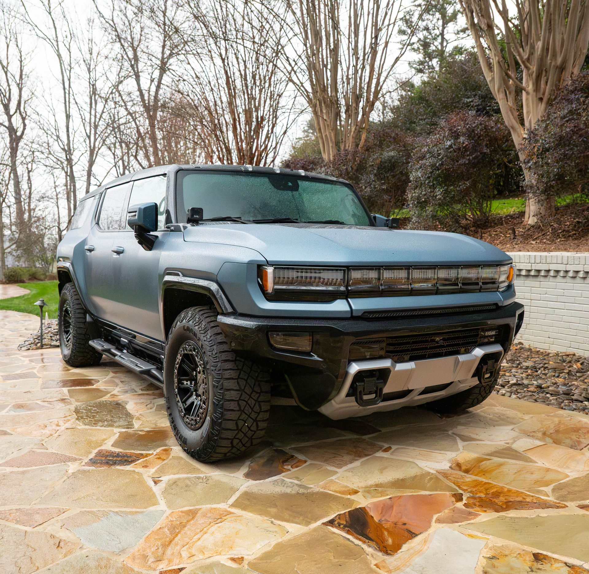 Blue Hummer EV SUV parked on a stone patio with a wooded background.