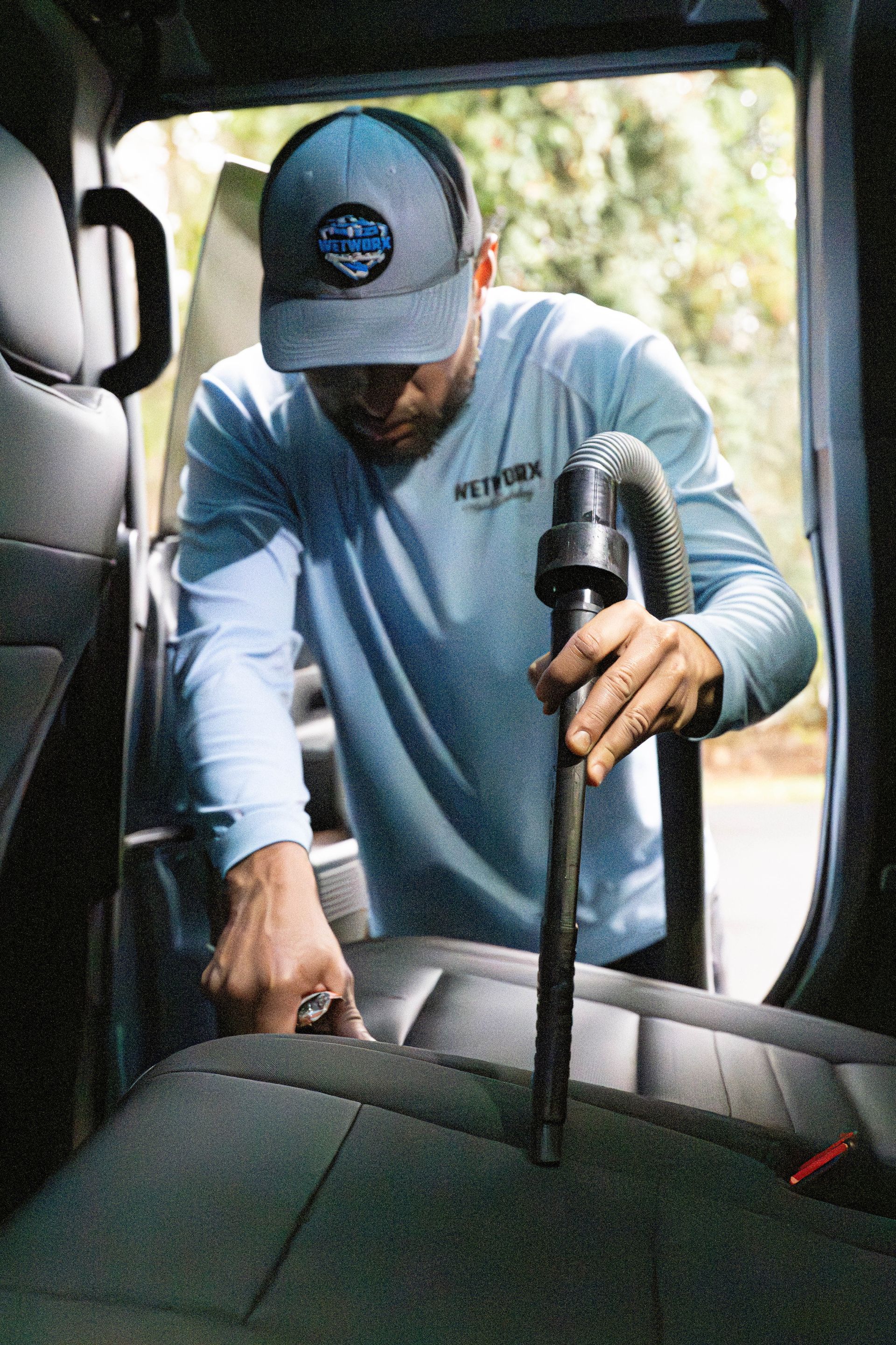 Man vacuuming a car seat inside a vehicle. He wears a blue shirt, cap, using a black vacuum.