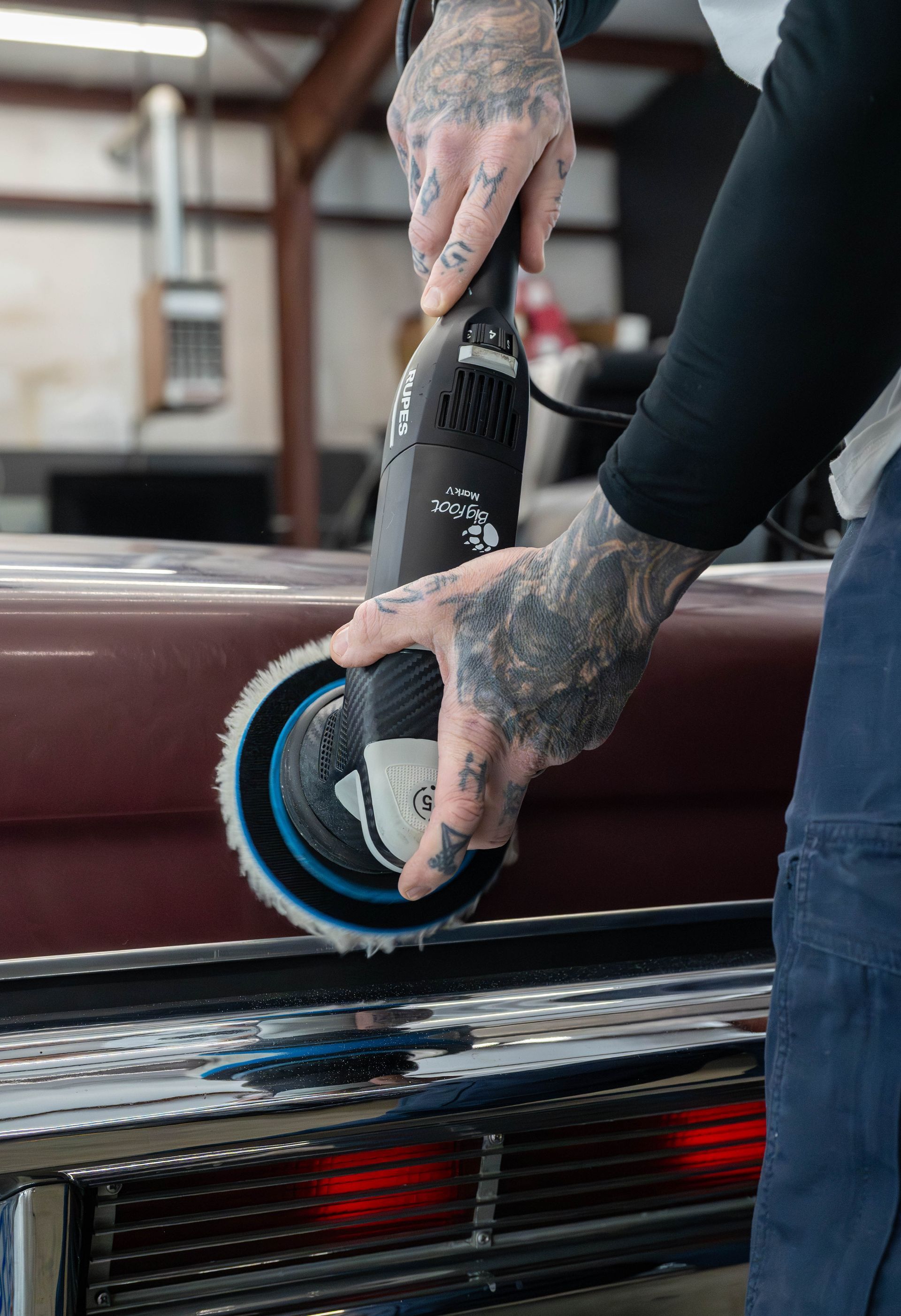 Person using a buffer to polish a car's bumper; visible tattoos, black sleeve.