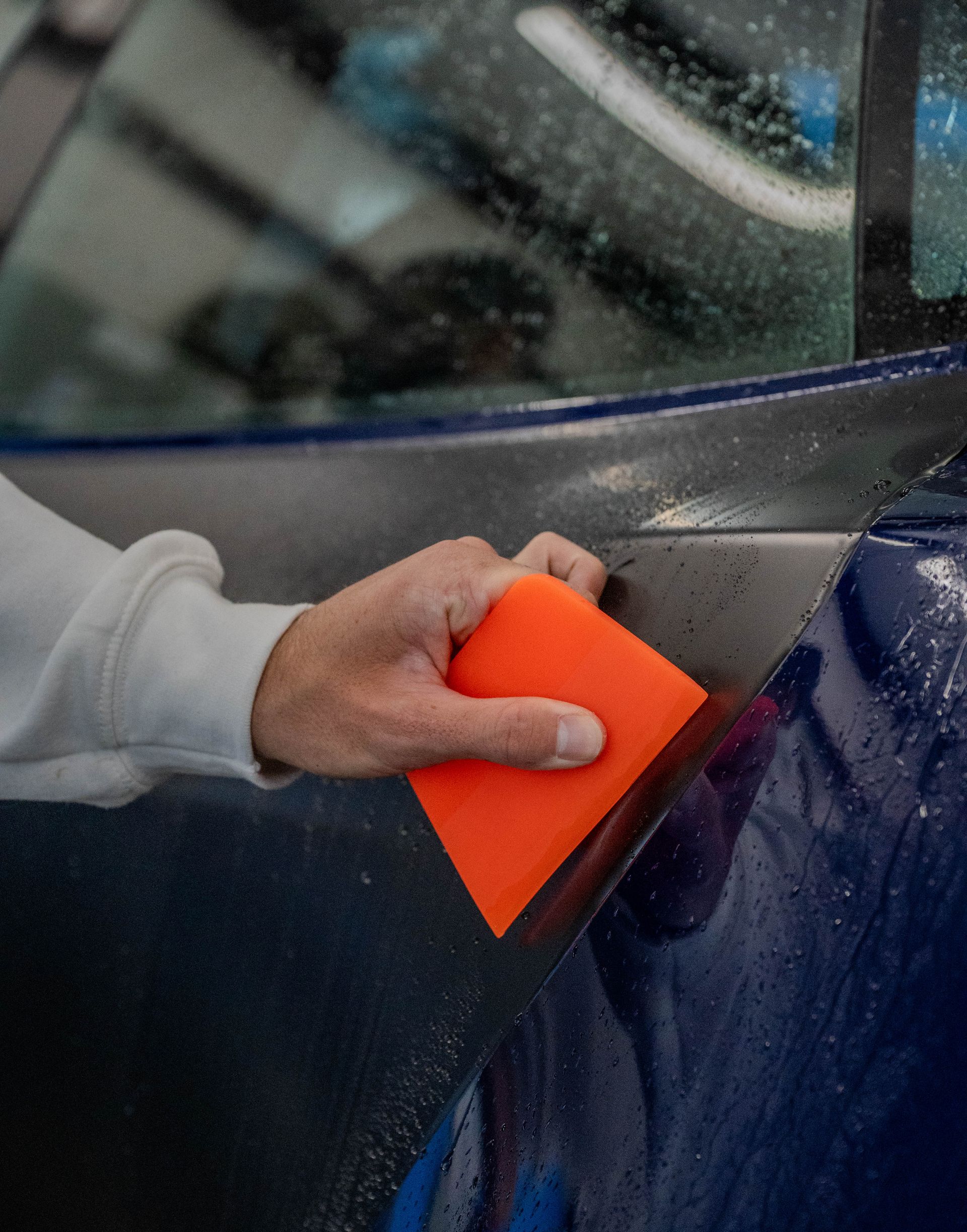 Person using an orange squeegee on a blue car door, likely applying a film.
