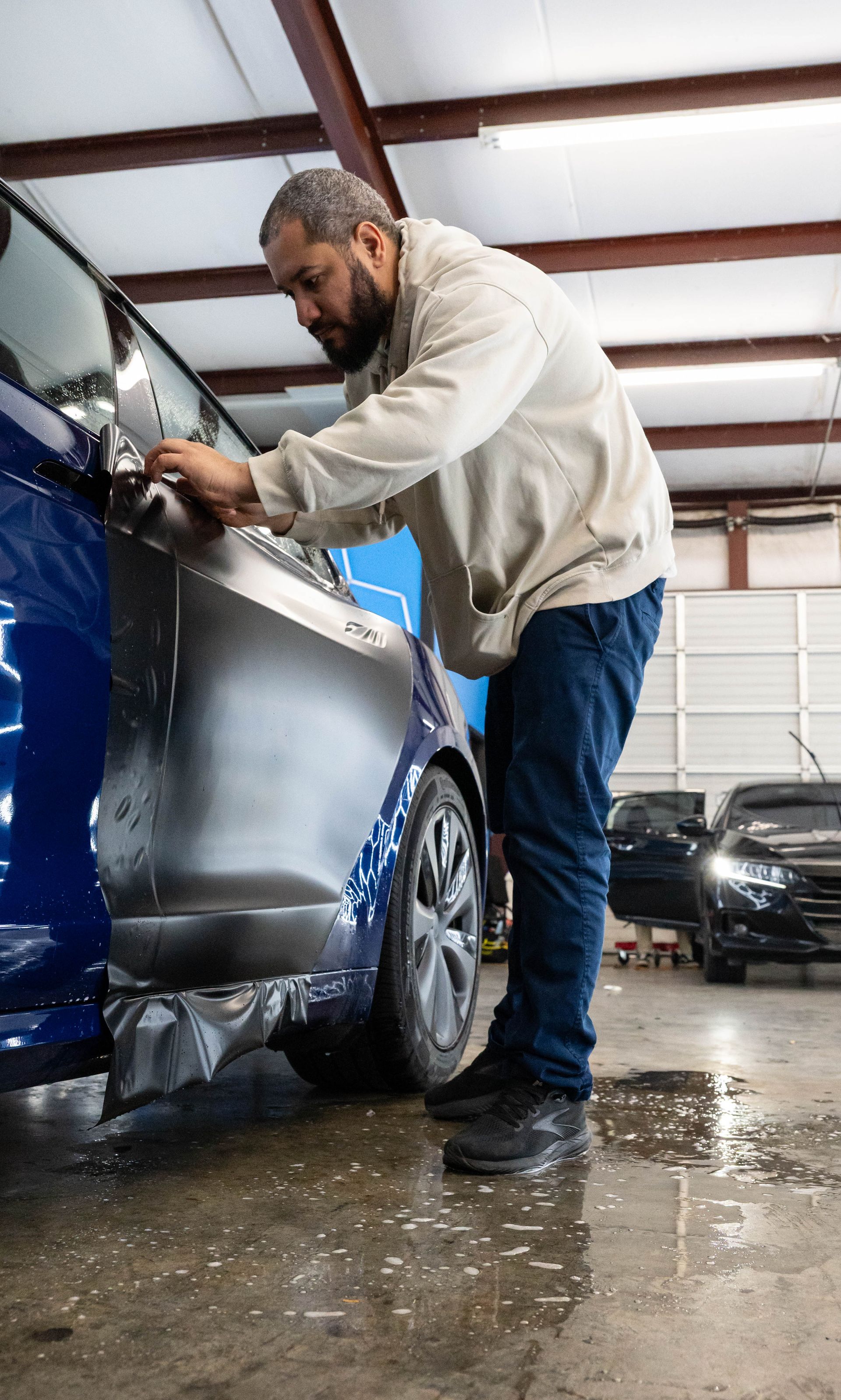 Man applying a dark tint film to a blue car window inside a shop.