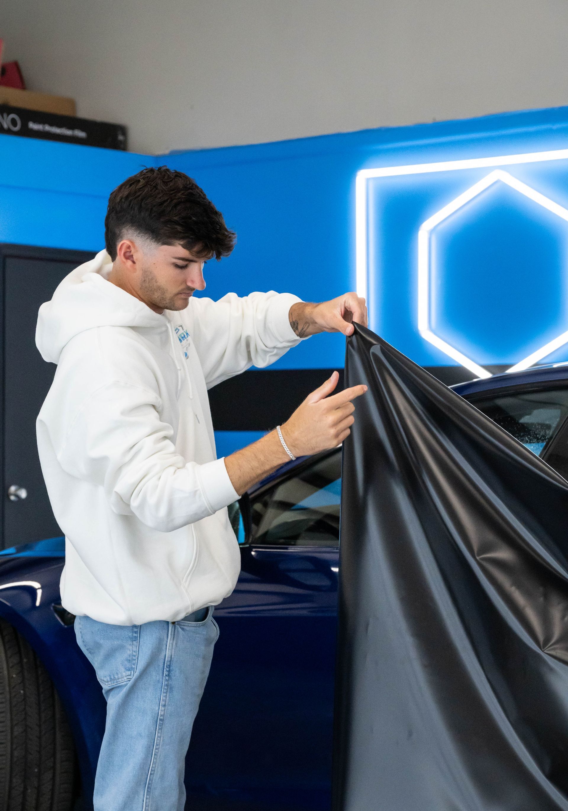 Person in white hoodie applying black vinyl wrap to a dark blue car in a garage.