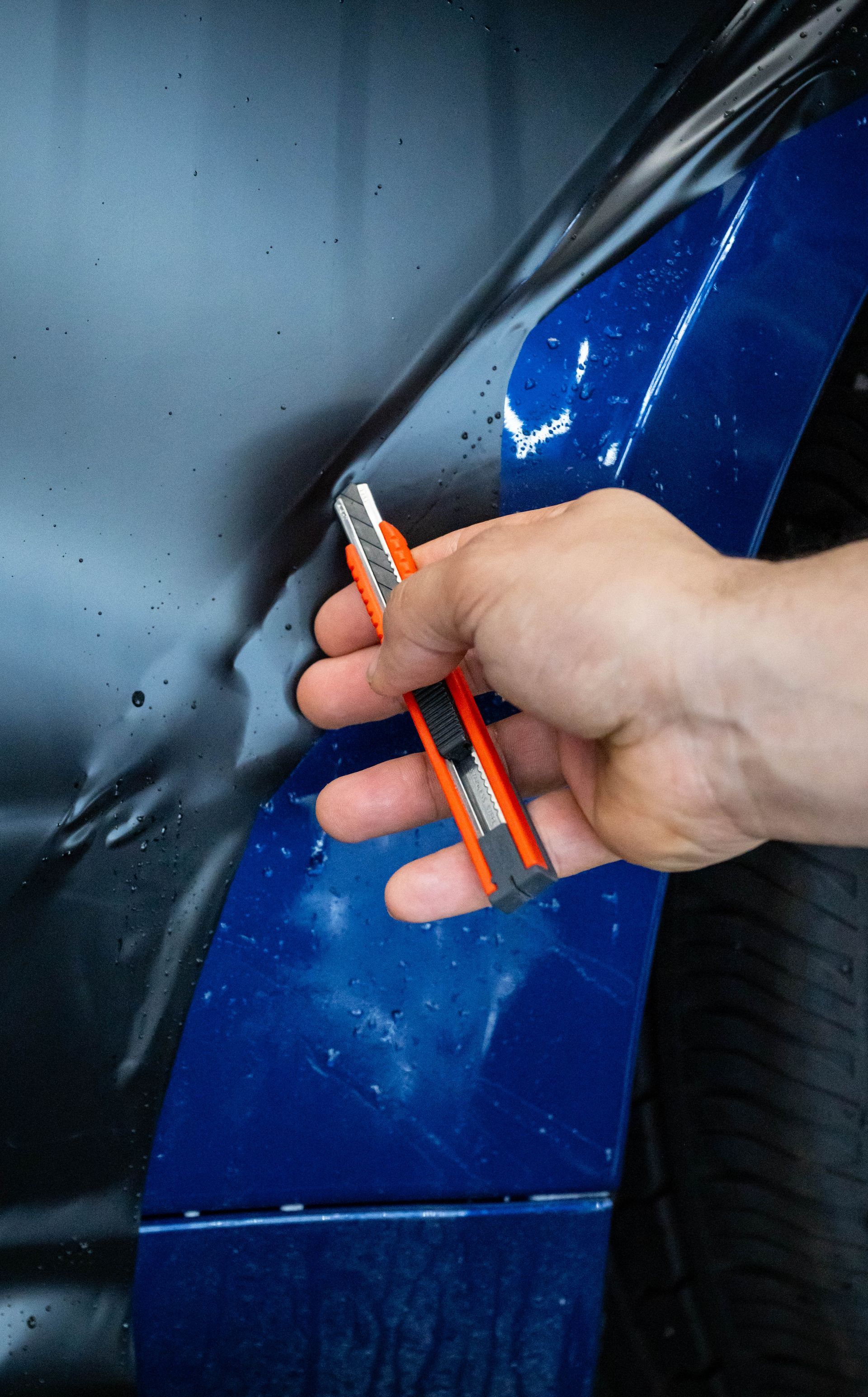 Hand cutting film on a blue car fender with an orange utility knife.