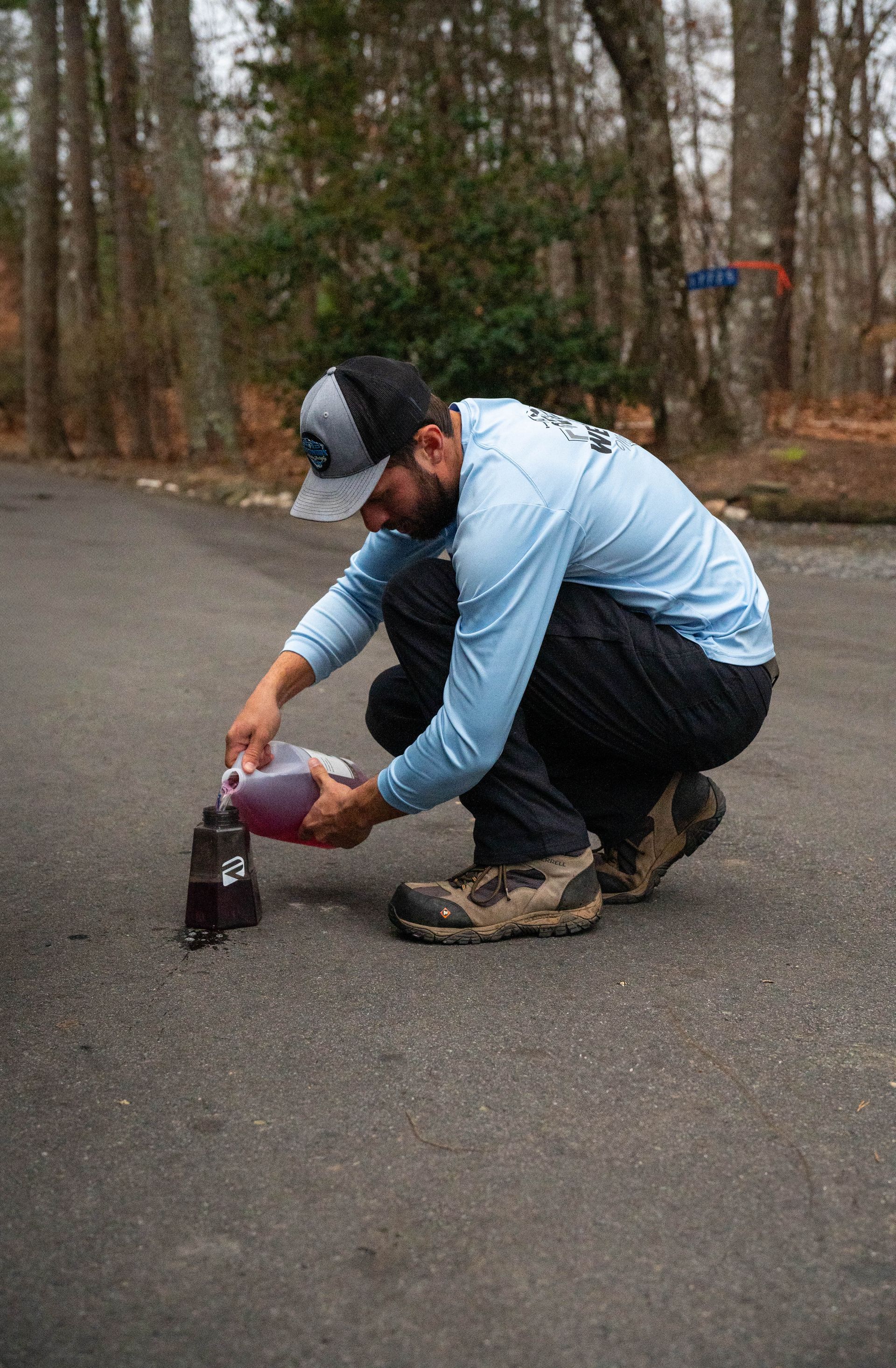 Man in blue shirt pours liquid from a container on a driveway.