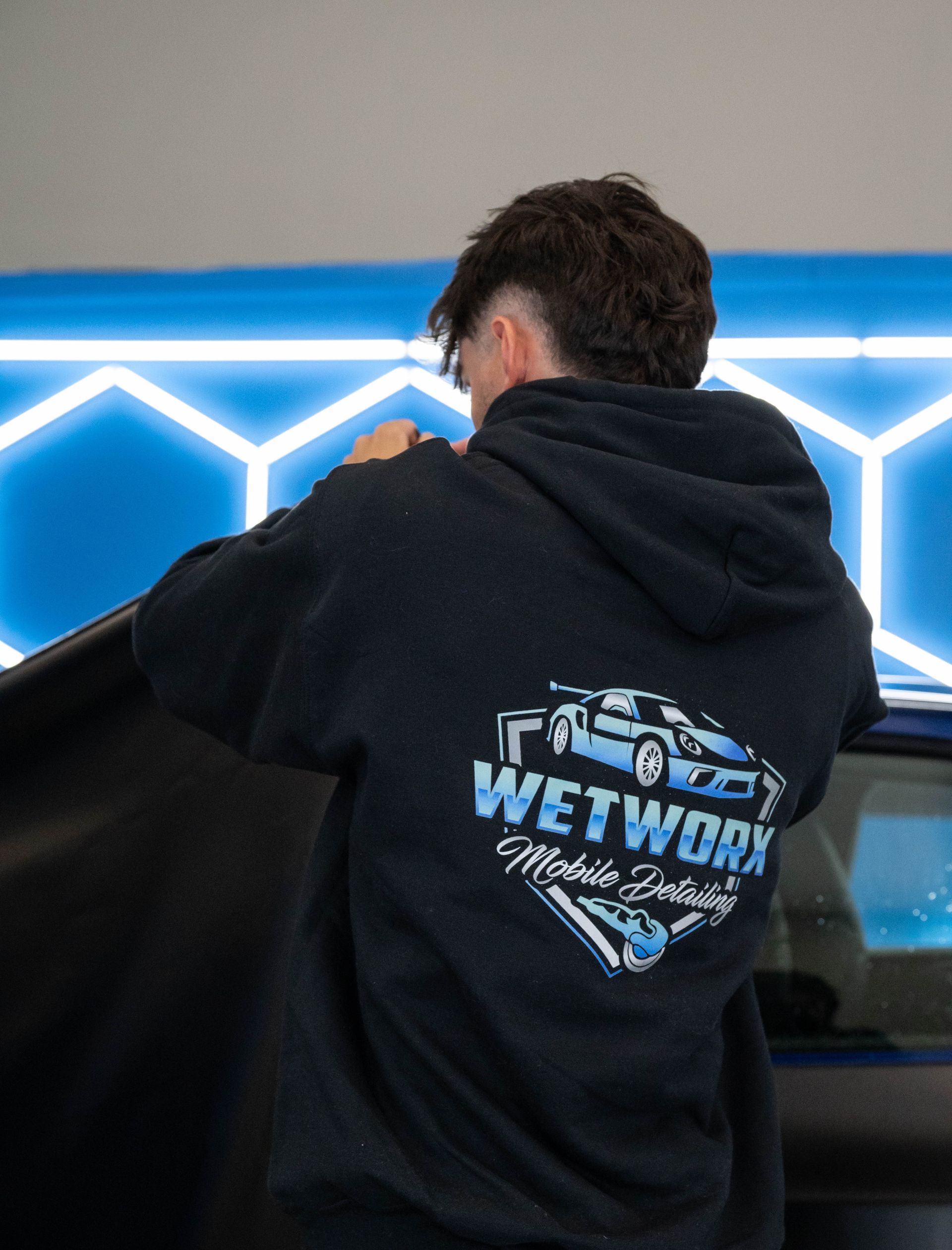 Person wearing a hoodie with Wetworx logo working on a car. Blue hexagon lights in the background.