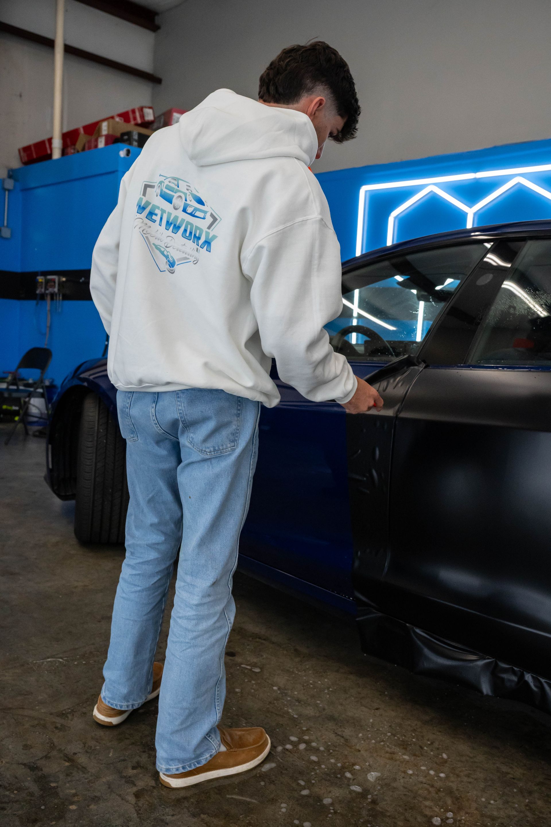 Person in a white hoodie inspecting a dark-colored car in a garage with neon blue lighting.