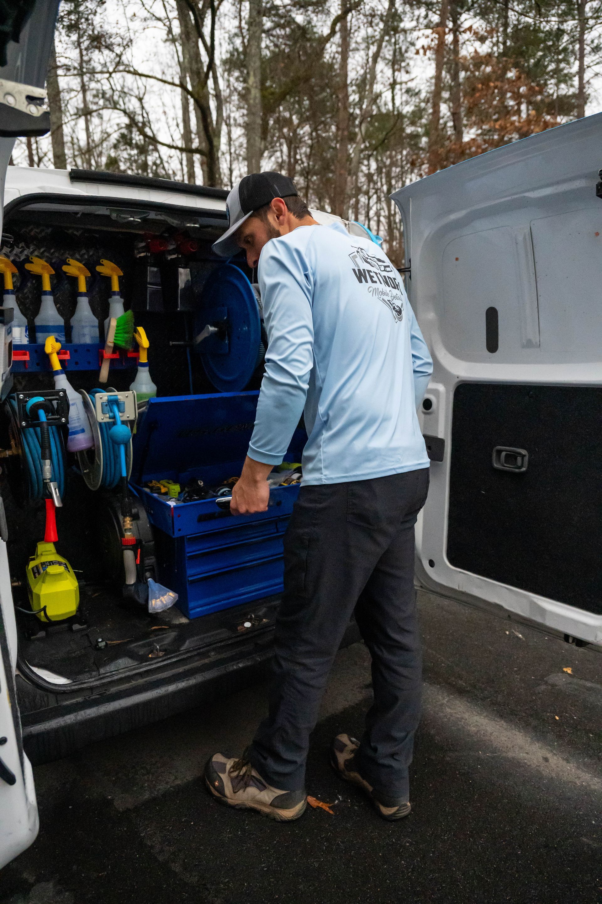Man washes car tire with a brush, outside. A white bucket and spray bottle are nearby.