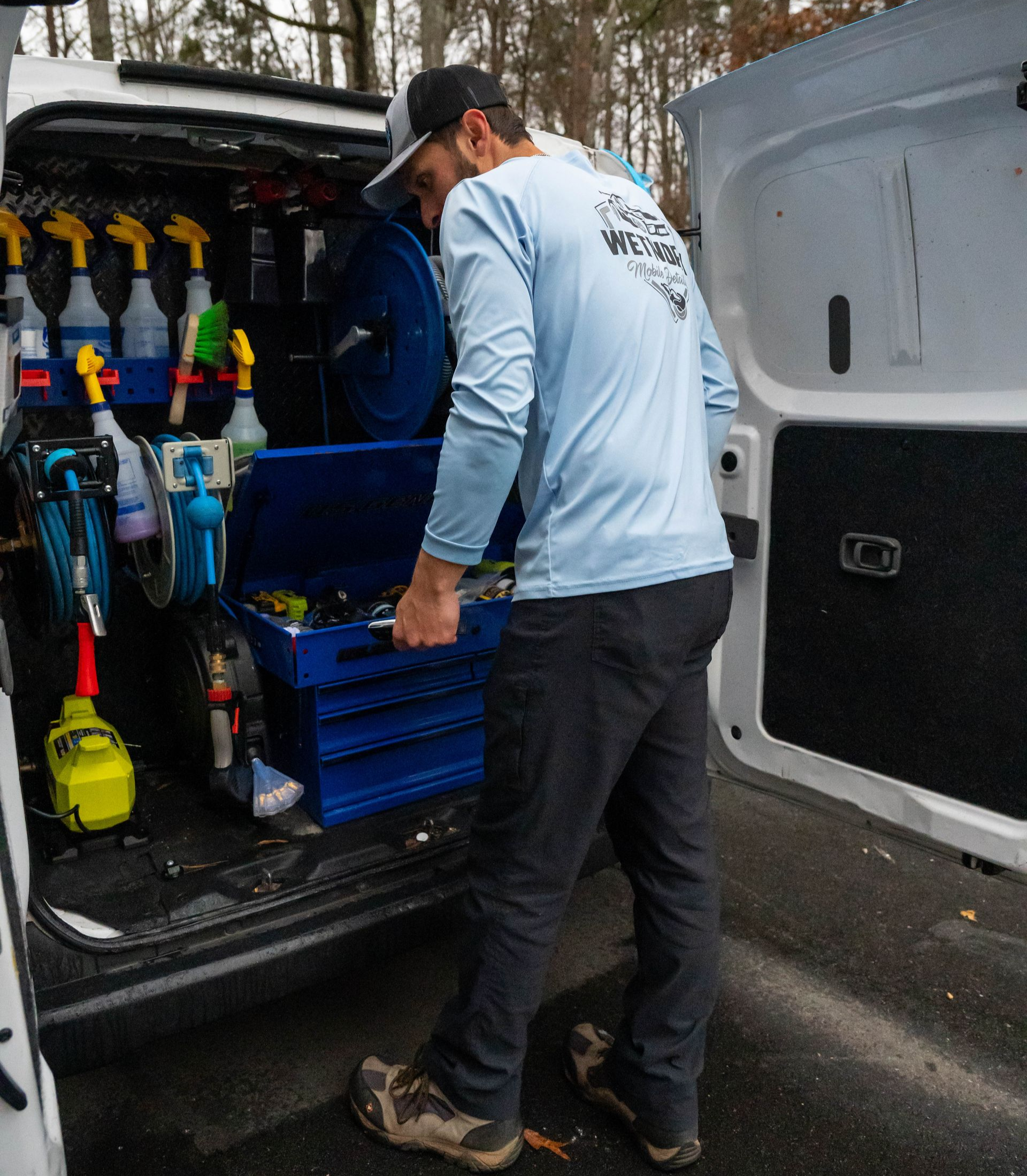 Man in light blue shirt removes toolbox from a van with spray bottles and tools inside.